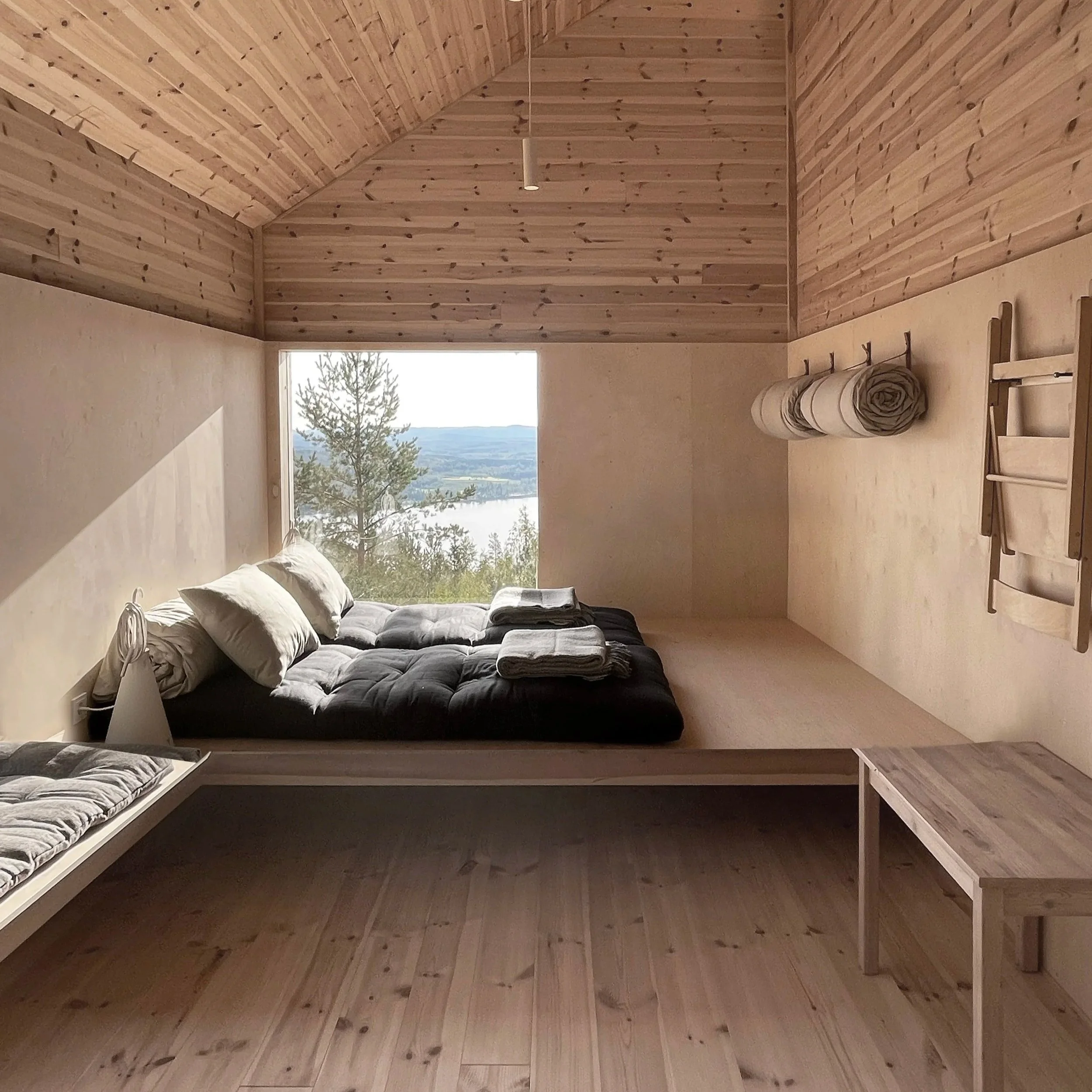 Minimalist bedroom with wooden walls and ceiling, a large window showing a scenic view of trees and water, a black bed with pillows and folded blankets, wall hooks with rolled-up mats, and a small wooden table.
