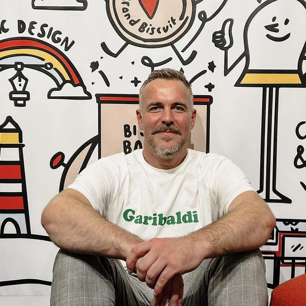 A man with short gray hair and a beard, wearing a white t-shirt with 'Garibaldi' written in green, sitting in front of a colorful, cartoon-style mural featuring various illustrations including a rainbow, a smiling character, and a coffee cup.