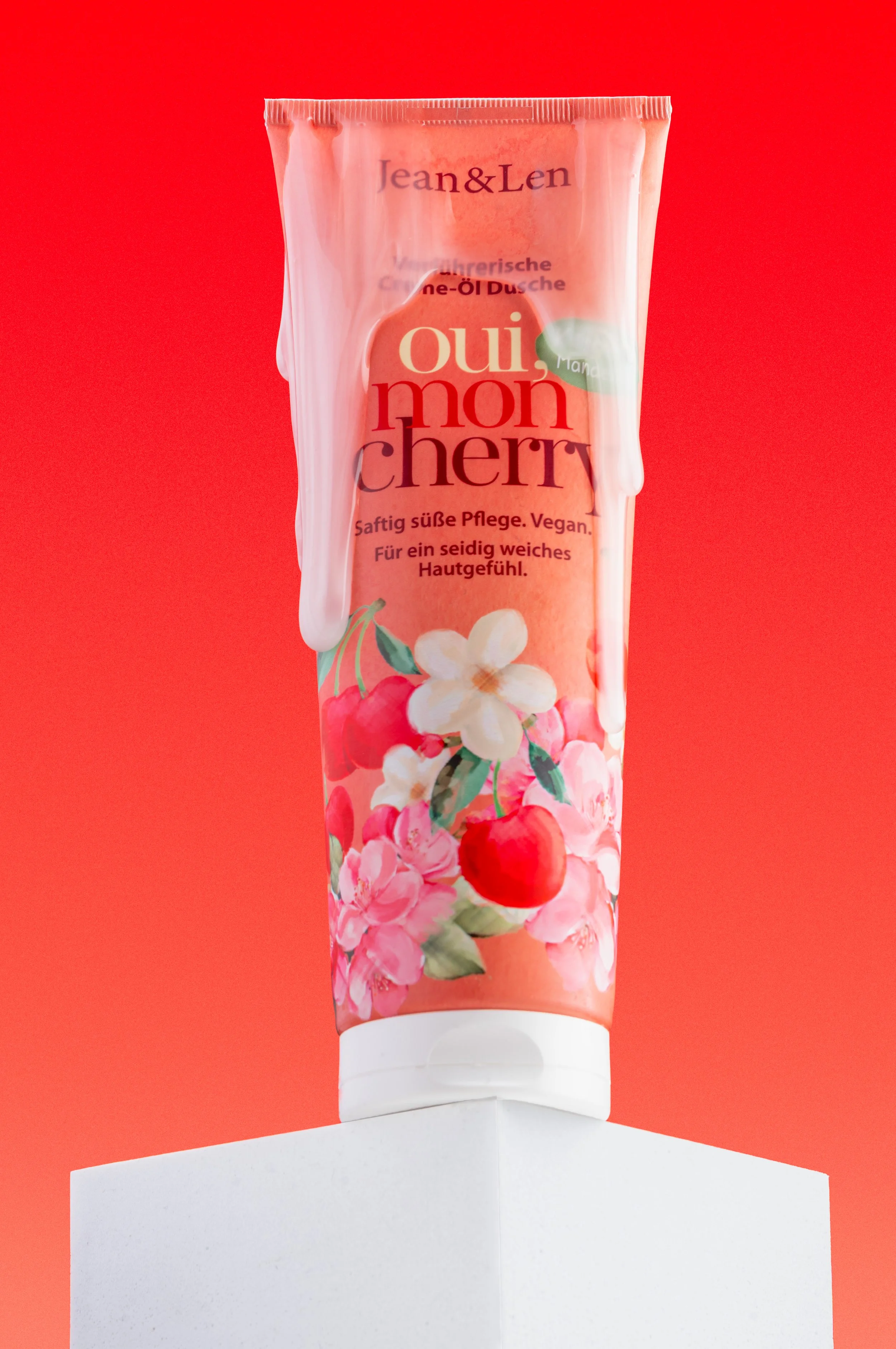 Kreative Studio-Produktfotografie der Jean & Len „Oui Mon Cherry“ Duschcreme mit tropfendem Creme-Effekt auf weißem Podest und rotem Hintergrund.