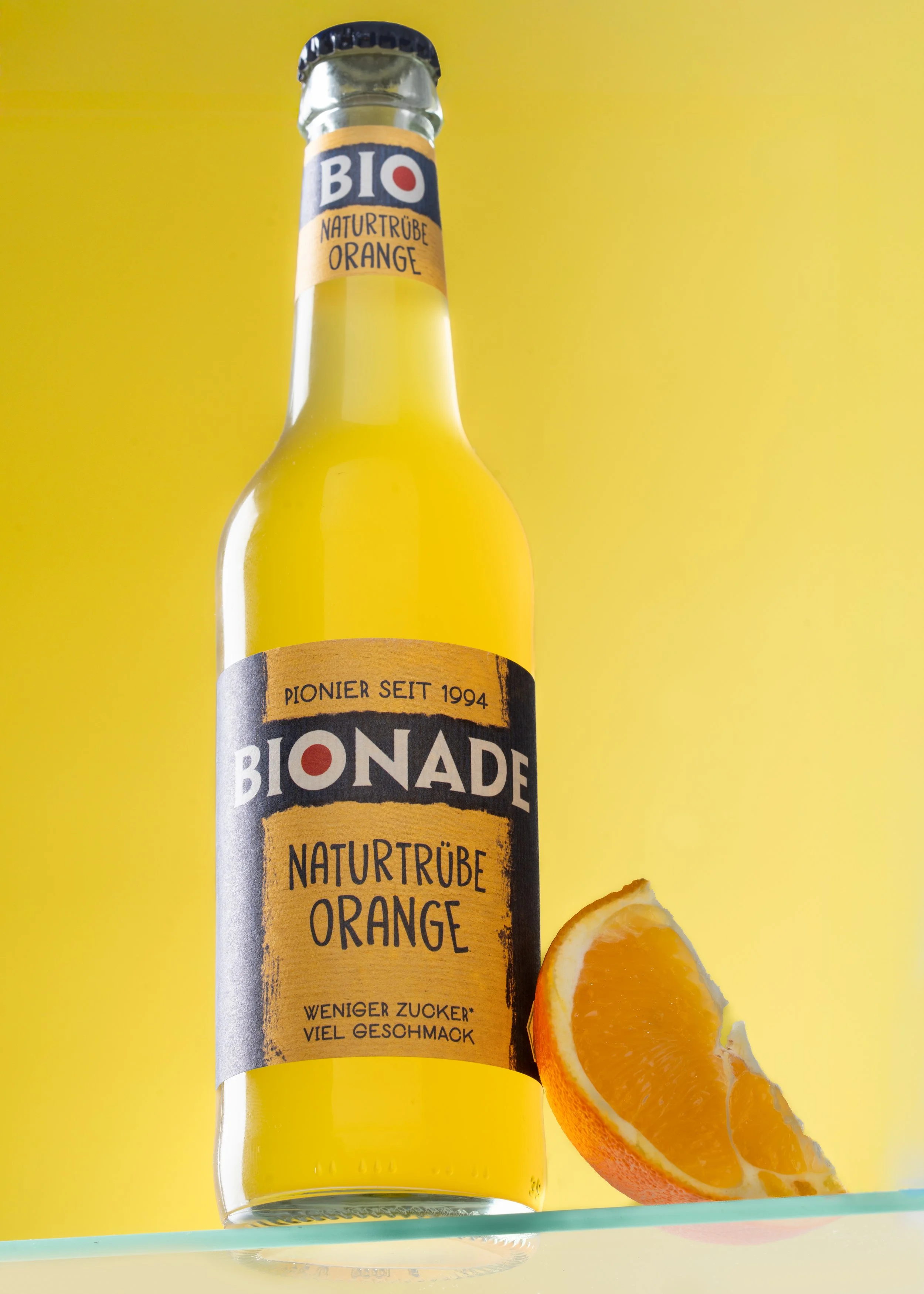 Studioaufnahme einer Bionade Orange Flasche mit Orangenscheibe vor gelbem Hintergrund