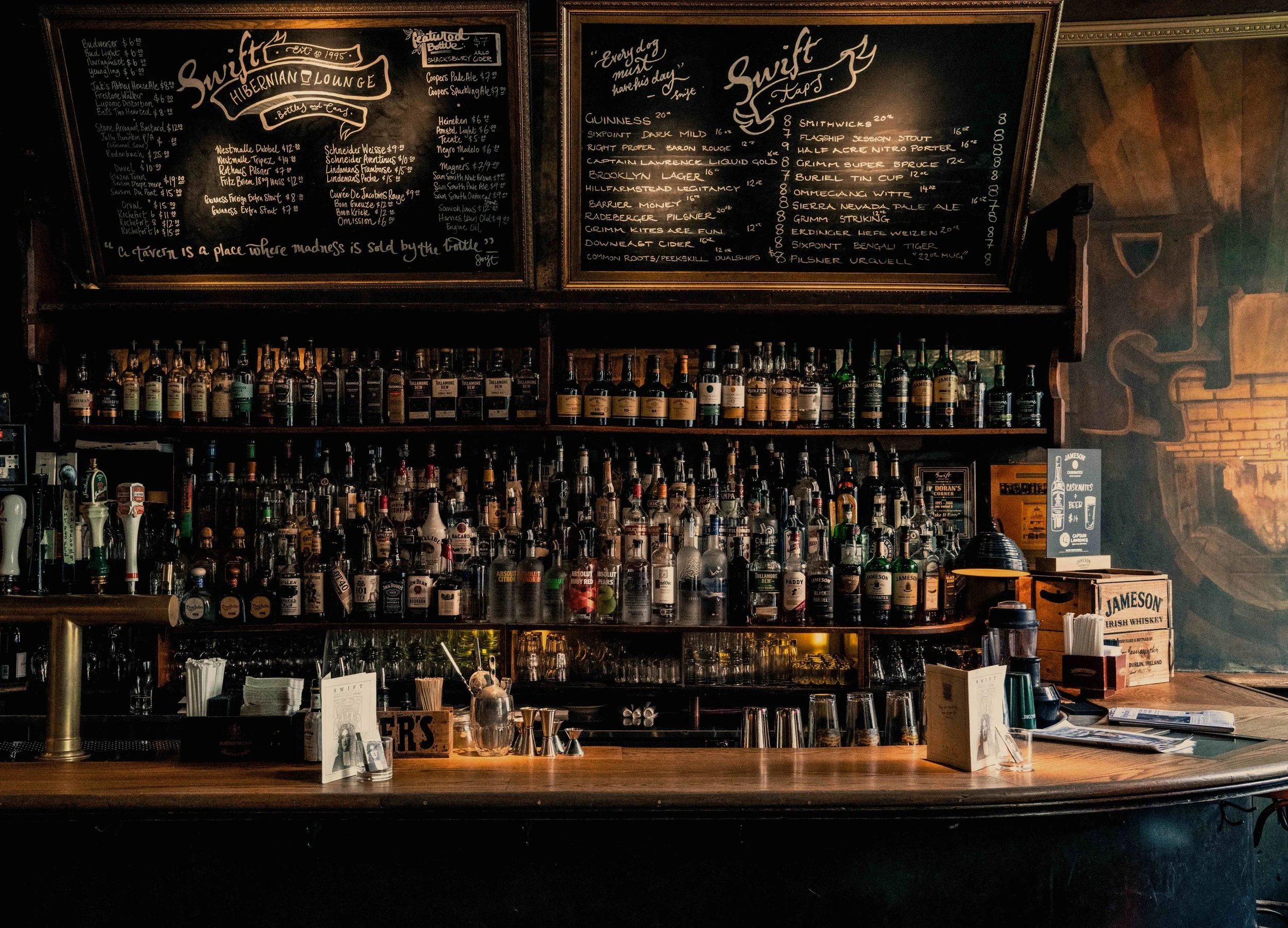 A bar with shelves filled with various liquor bottles, a wooden counter with glasses, napkins, and a menu. Two chalkboard menus with handwritten drink options and prices are displayed above the shelves, and there is a mural of a fireplace on the wall to the right.