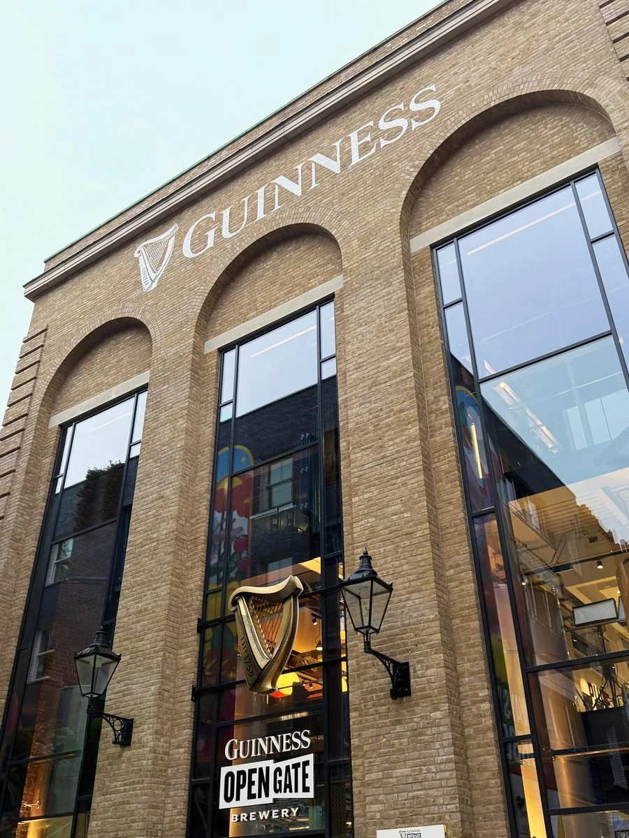 Exterior of a Guinness brewery building with a large Guinness logo, stained glass windows, and an open gate sign.