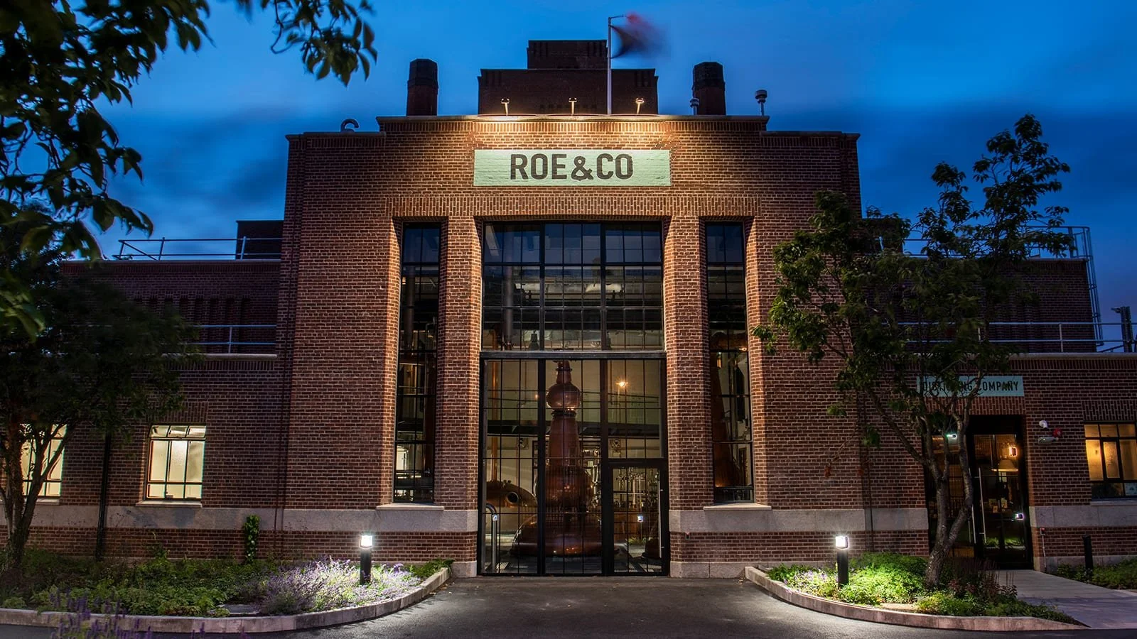 A brick building with large windows, a sign that reads 'ROE&CO,' and a large copper still inside, illuminated at night with surrounding trees and landscaped areas.