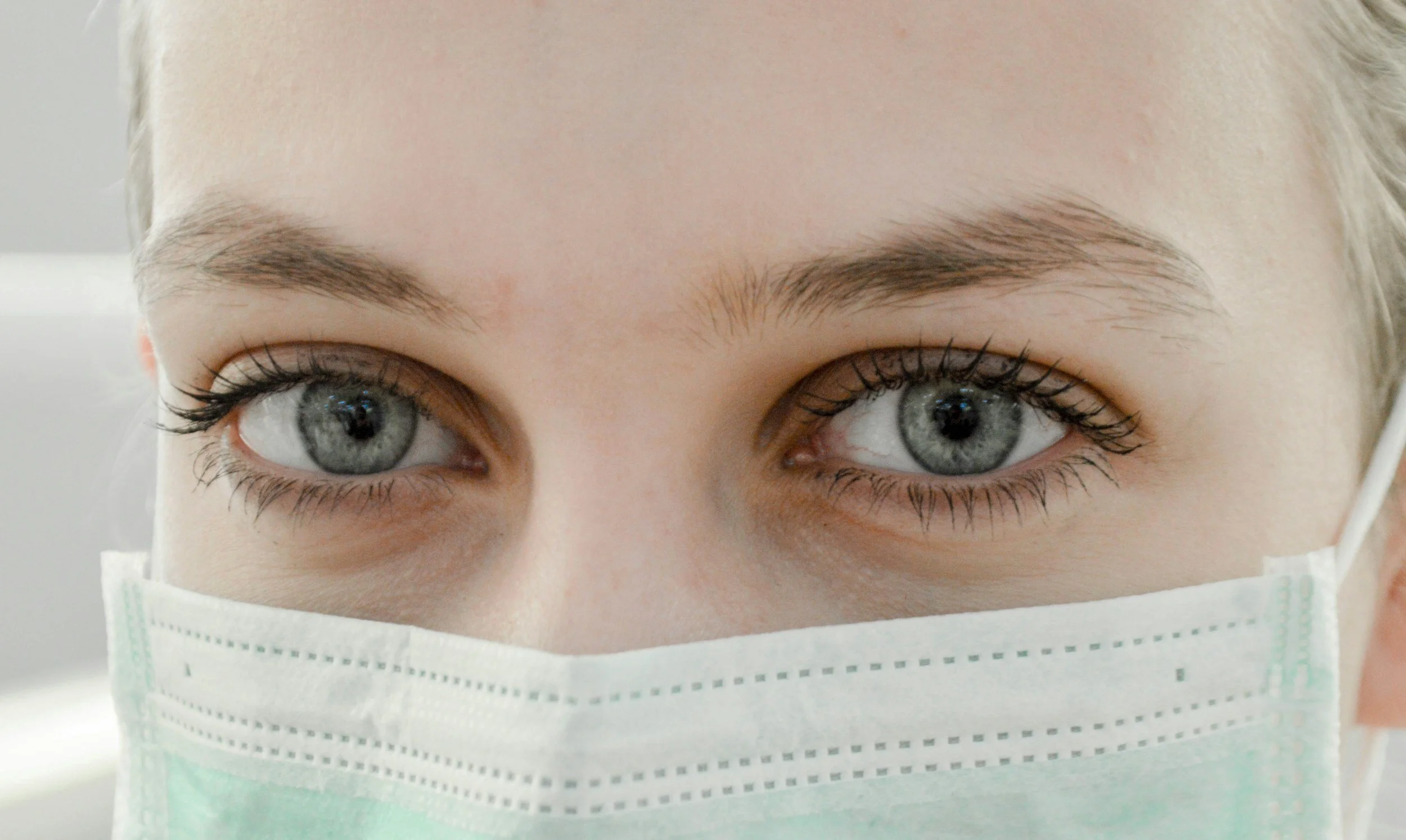 Close-up of a person's face with light skin, blue eyes, and dark eyelashes, wearing a medical face mask, against a neutral background. Therapy for Healthcare Providers.