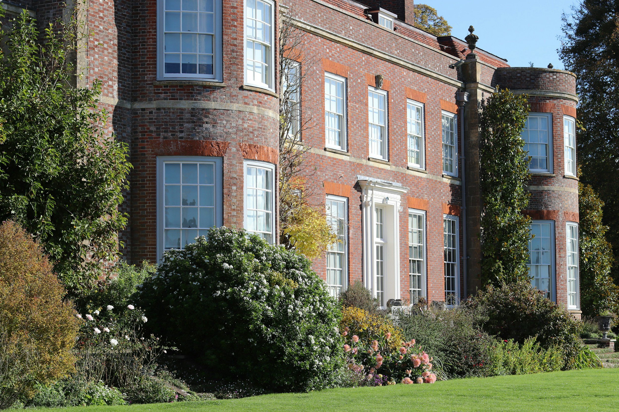 A large brick mansion with multiple large windows, surrounded by well-maintained garden with various shrubs, flowering plants, and a lush green lawn.