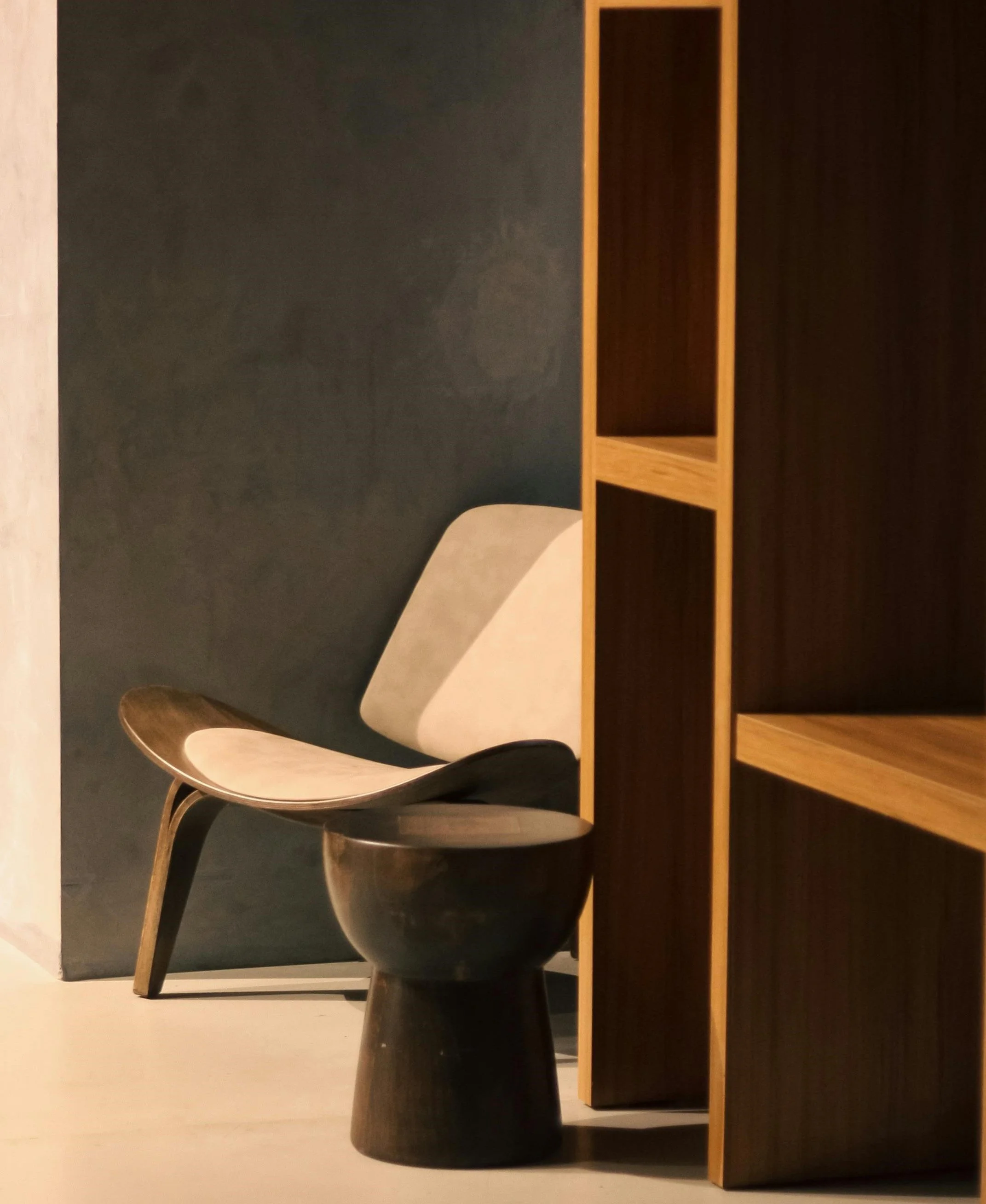 A modern interior with a wooden shelf, an abstract chair with curved wood and fabric, and a dark wall in the background.