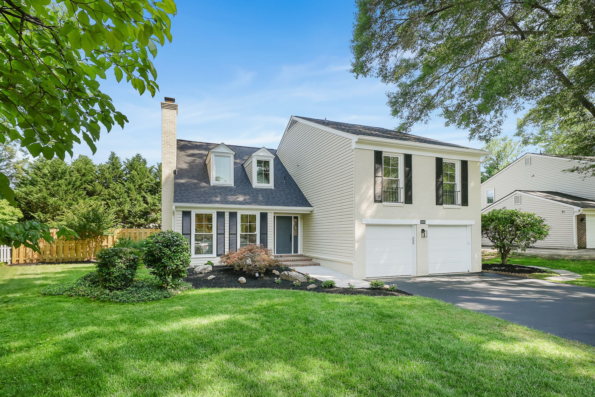 A two-story house with white siding and black shutters, a black roof, two dormer windows, a front door with steps, a double garage, and a well-maintained lawn with bushes and trees.