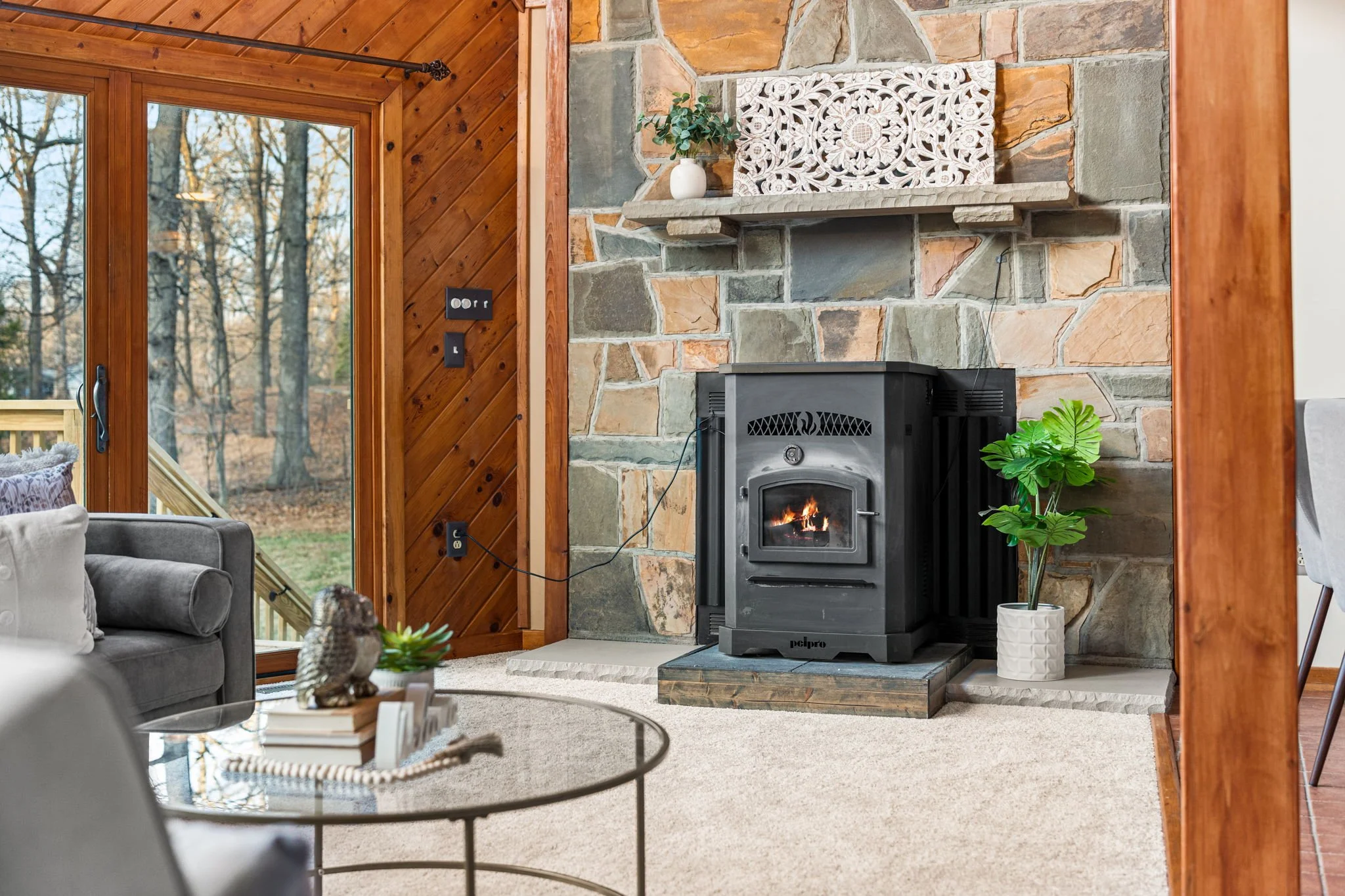 Living room with a stone fireplace, black stove with a small fire, and decorative plants, with wooden beams and large glass doors leading outside.
