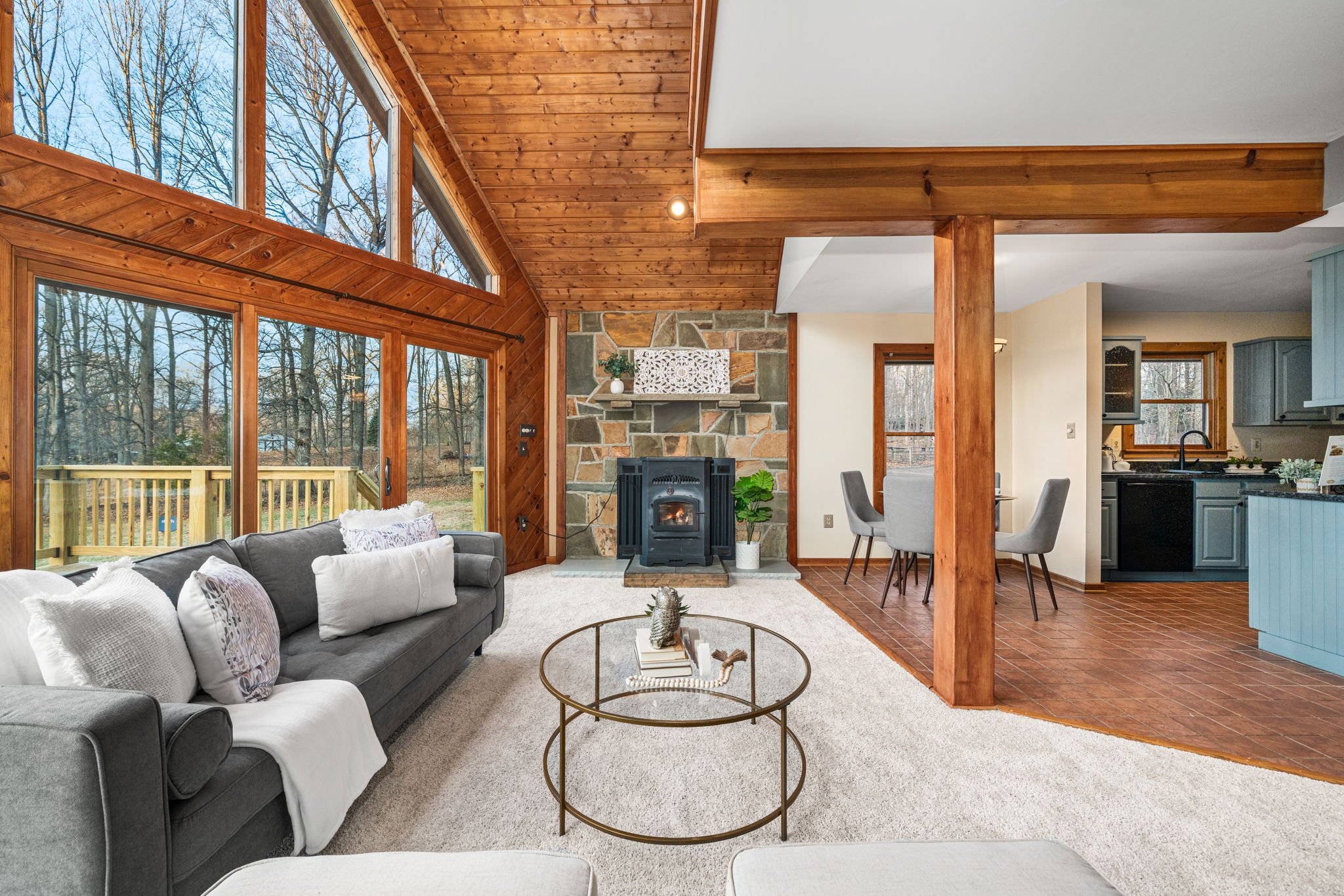 Living room with large windows, gray sofas with white pillows, a round coffee table, a stone fireplace, and a view of a backyard with trees.