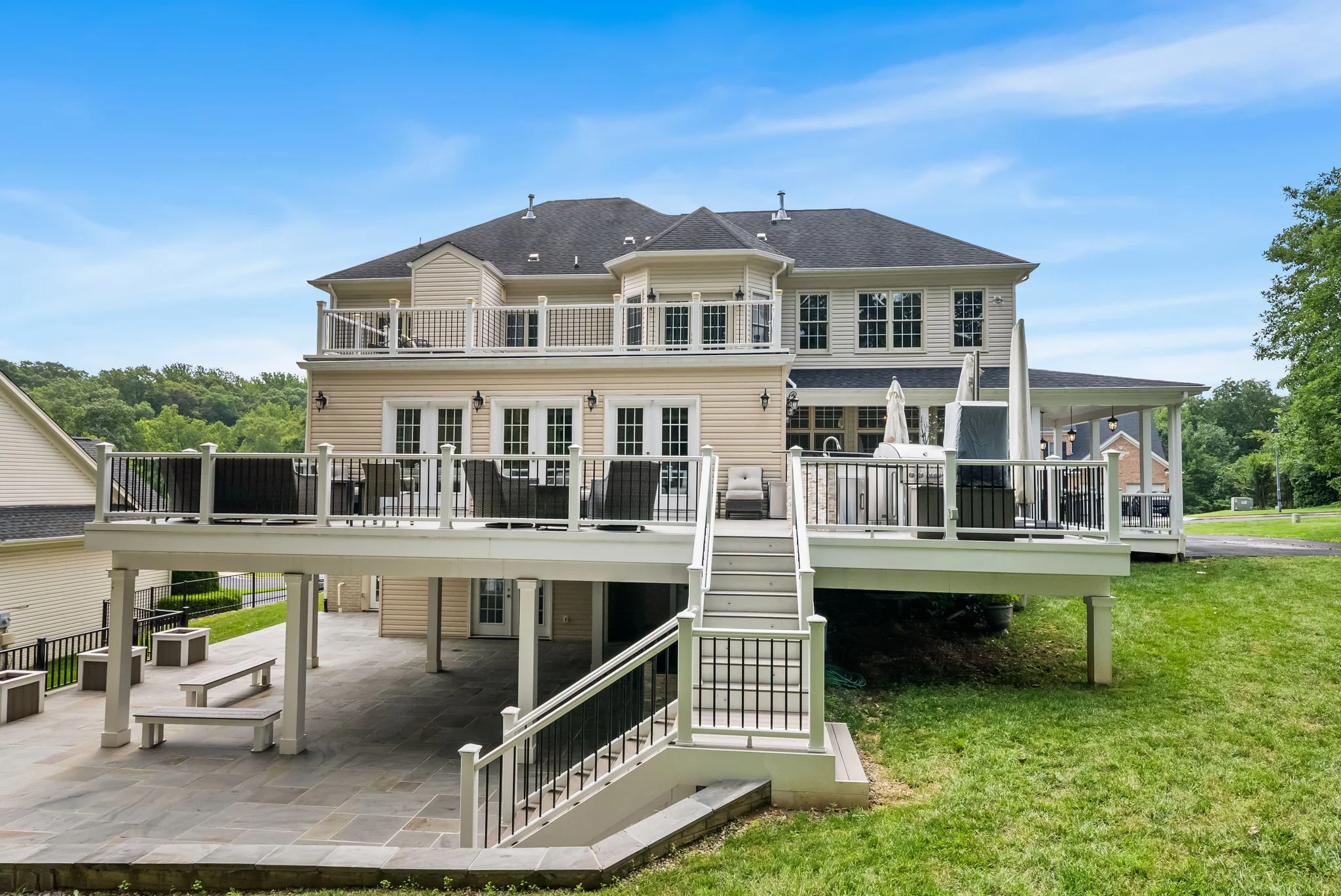 Large two-story house with multiple decks and stairs, beige siding, black railings, outdoor patio with benches, and a landscaped yard with green grass and trees.