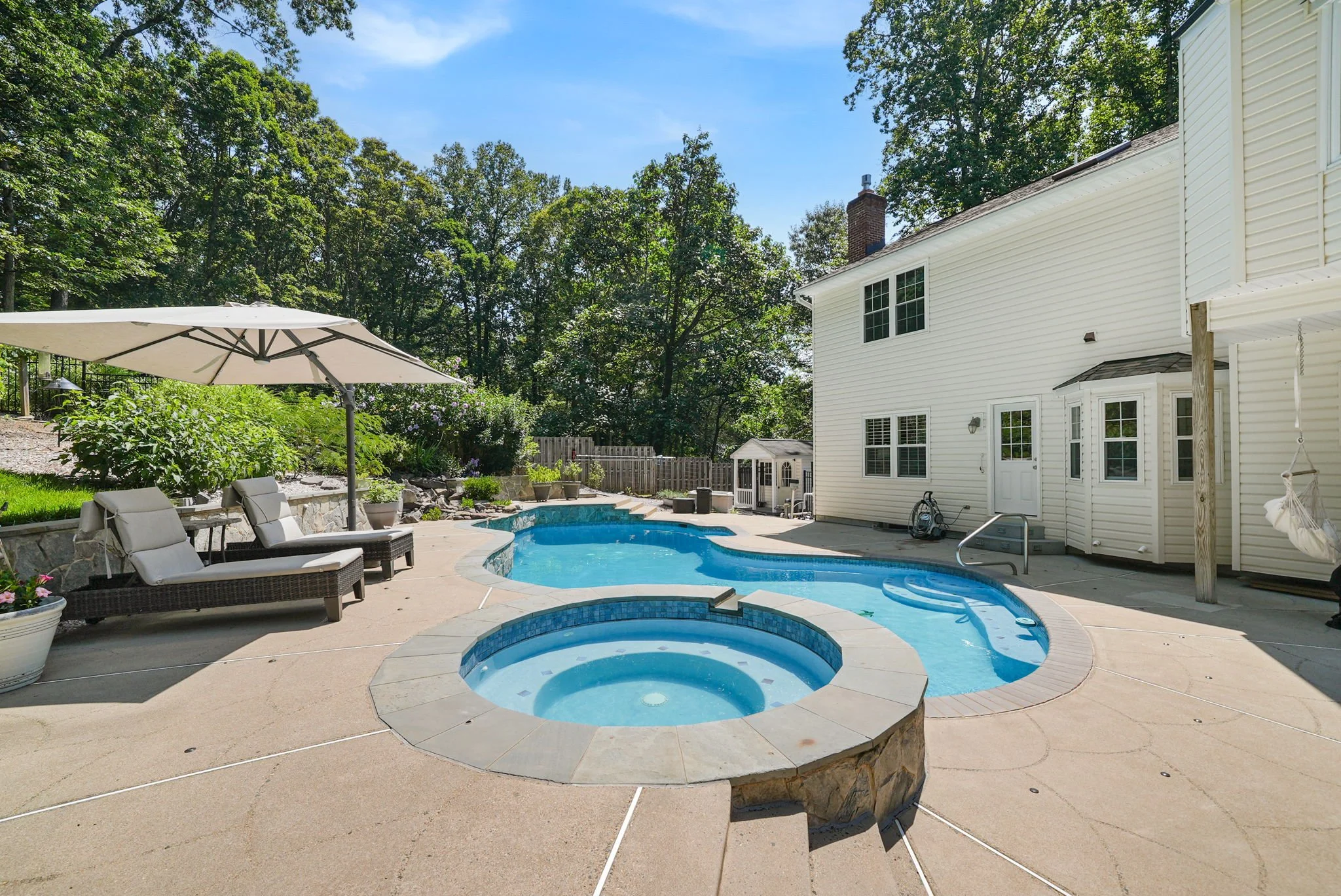 Backyard with swimming pool, hot tub, lounge chairs, patio umbrella, and playhouse, surrounded by trees.