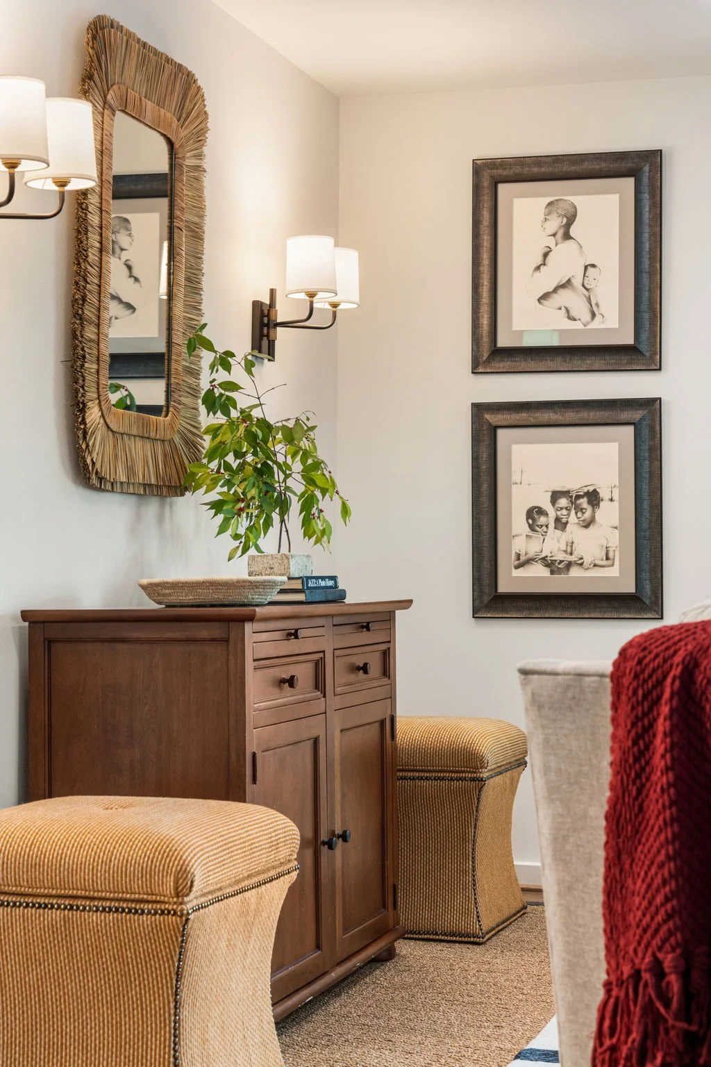 Living room corner with a wooden sideboard, a potted plant, two framed black and white photographs on the wall, a mirror with a woven frame, two wall lamps, and a beige upholstered chair with a red blanket.