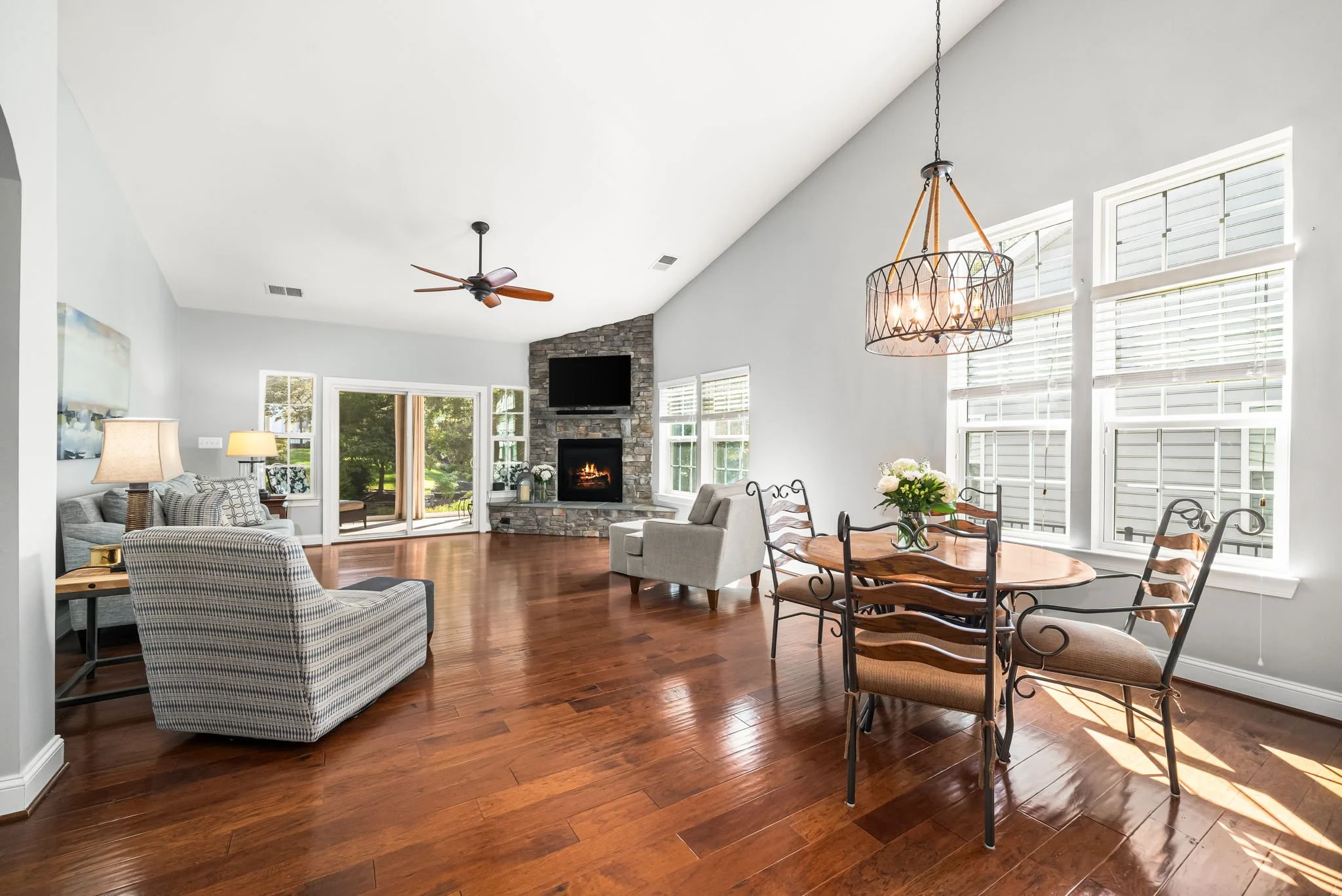 Bright open living room with hardwood floors, gray walls, large windows, a stone fireplace, a wall-mounted TV, a gray armchair, a checkered armchair, a sofa, a ceiling fan, a sliding glass door leading outside, and a dining table with chairs and a fl