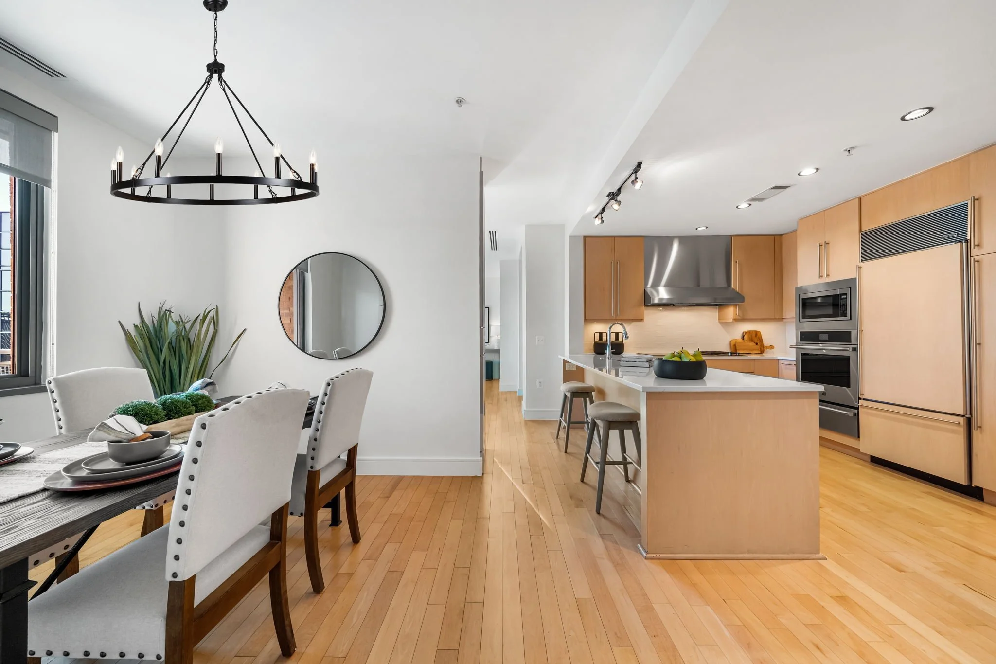 Modern kitchen and dining area with light wood floors, white walls, a circular chandelier above the dining table, and a kitchen island with barstools.