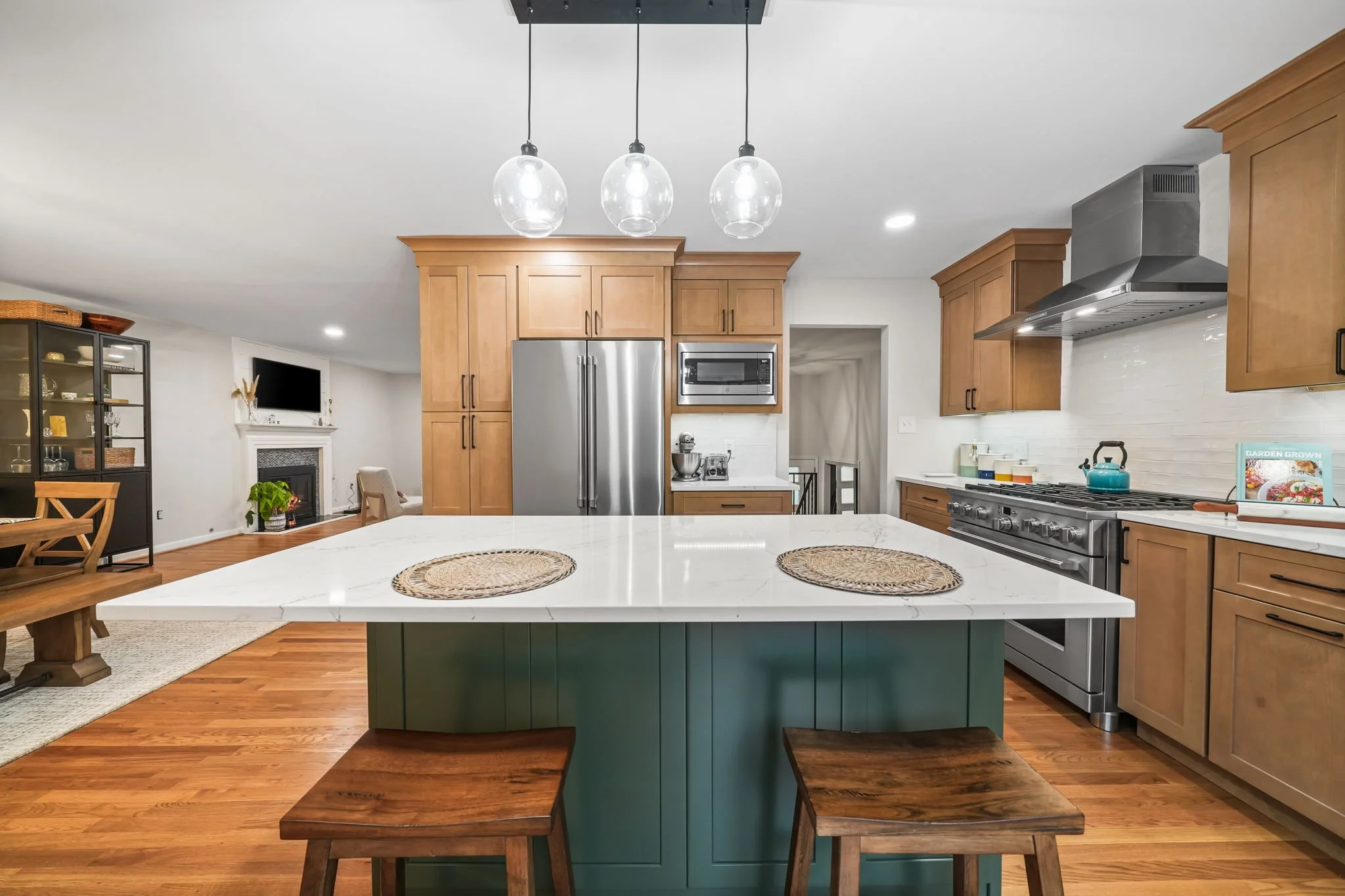 Modern kitchen with wooden cabinets, stainless steel refrigerator, oven, and microwave. White marble countertop island with two woven placemats, wooden stools, and a view of the living room with a fireplace and TV in the background.