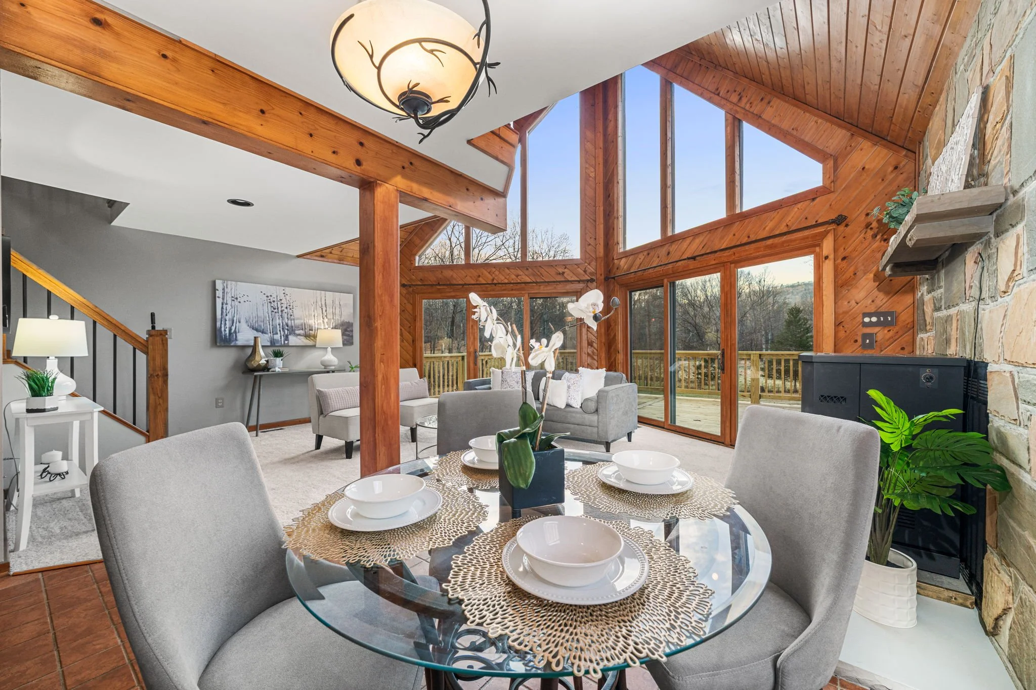Open-concept living and dining area with large windows and wood accents, featuring a glass dining table, grey chairs, and a grey sofa, decorated with plants and art.