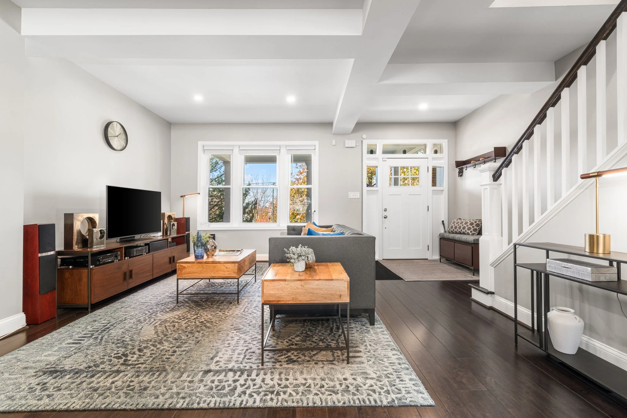 Living room with modern decor, dark hardwood floors, gray couch, wooden coffee tables, large window views, white walls, and a staircase with white balustrade.