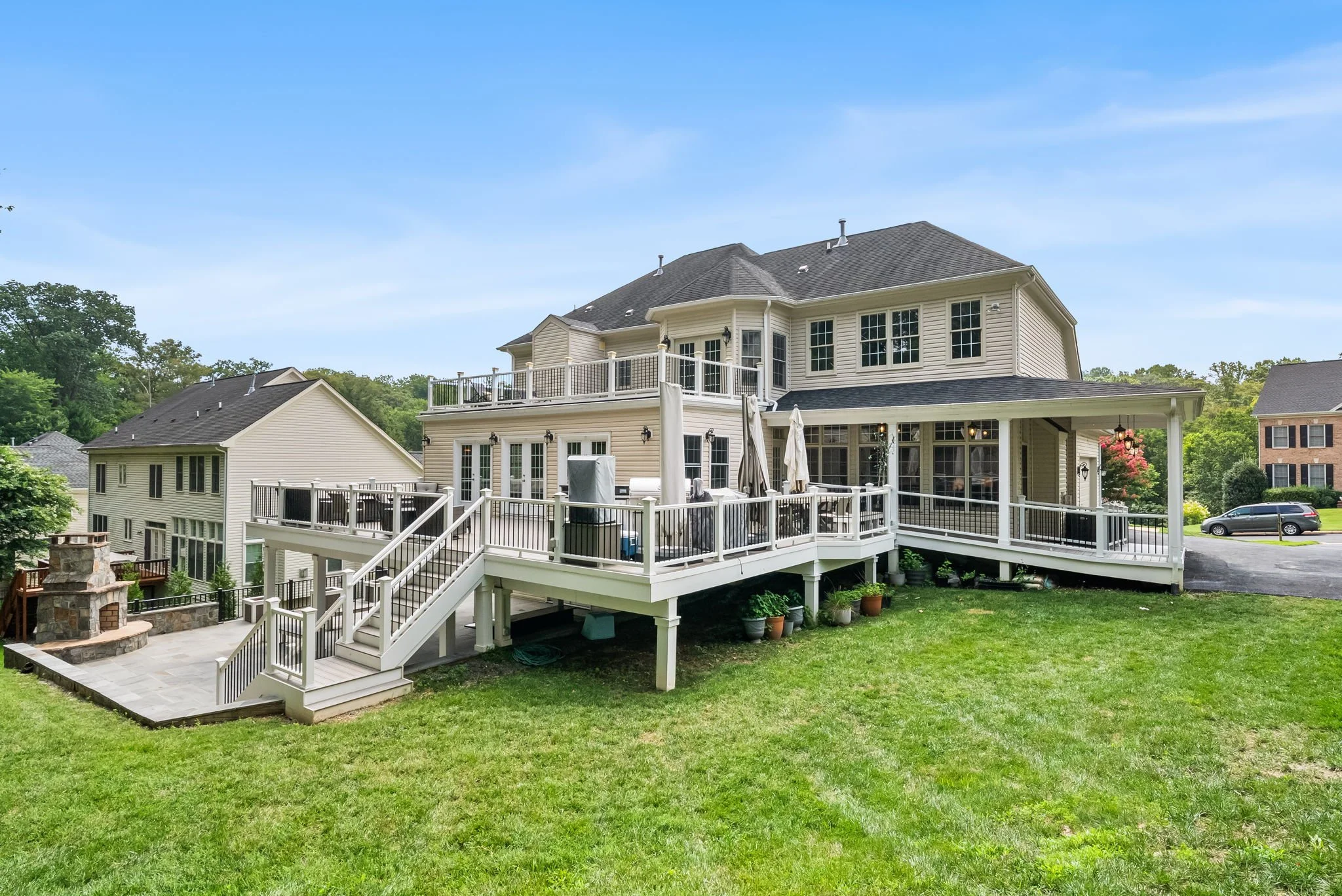 Large beige house with multiple decks and a staircase, surrounded by green grass, in a suburban neighborhood.
