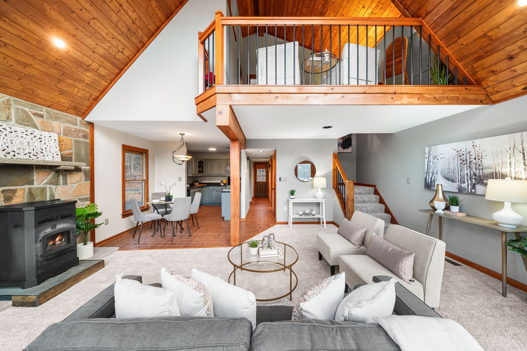 Spacious living room with a stone fireplace, beige and white sofas, and a glass coffee table. An open dining area with a table and chairs, adjacent to a kitchen with blue cabinets. A staircase leading to a loft with a wooden railing. Decor includes l