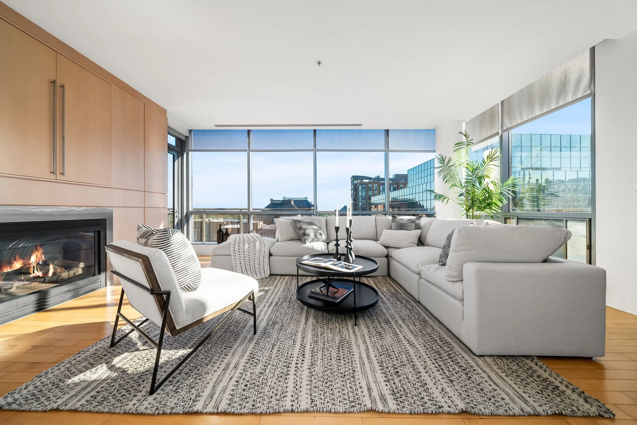 Modern living room with large windows, white sectional sofa, black coffee tables, armchair, fireplace, wooden floor, and a rug, with cityscape view outside.