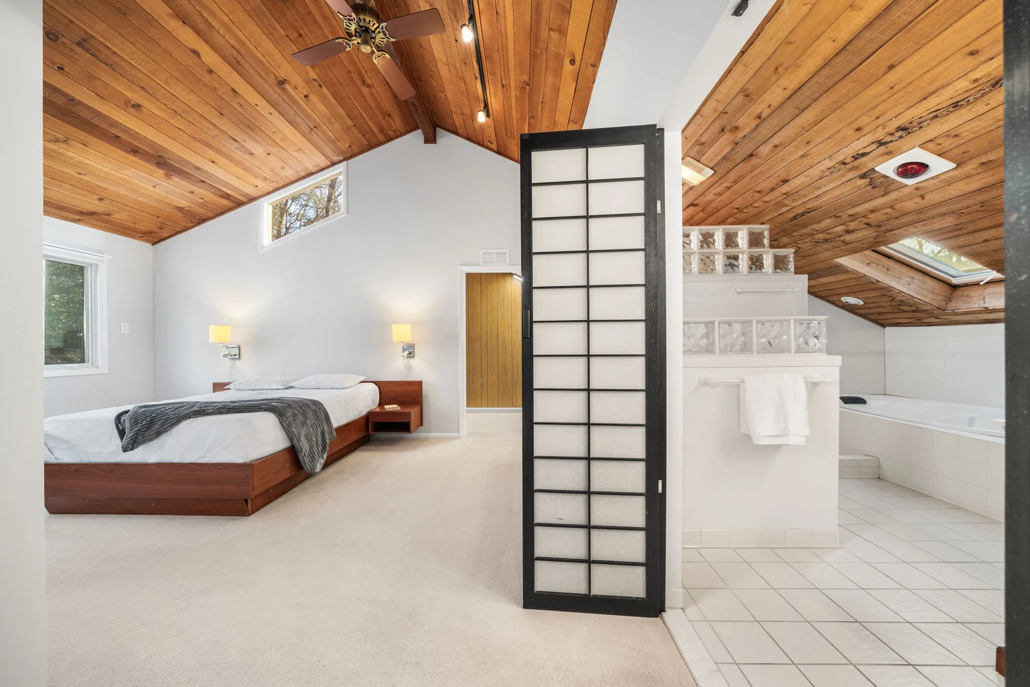 A bedroom with white walls and a wooden ceiling, a bed with white linens and a gray throw, two wall-mounted lamps, a small bedside table, and a partially open black folding screen leading to a bathroom with a bathtub, towels, and skylights.