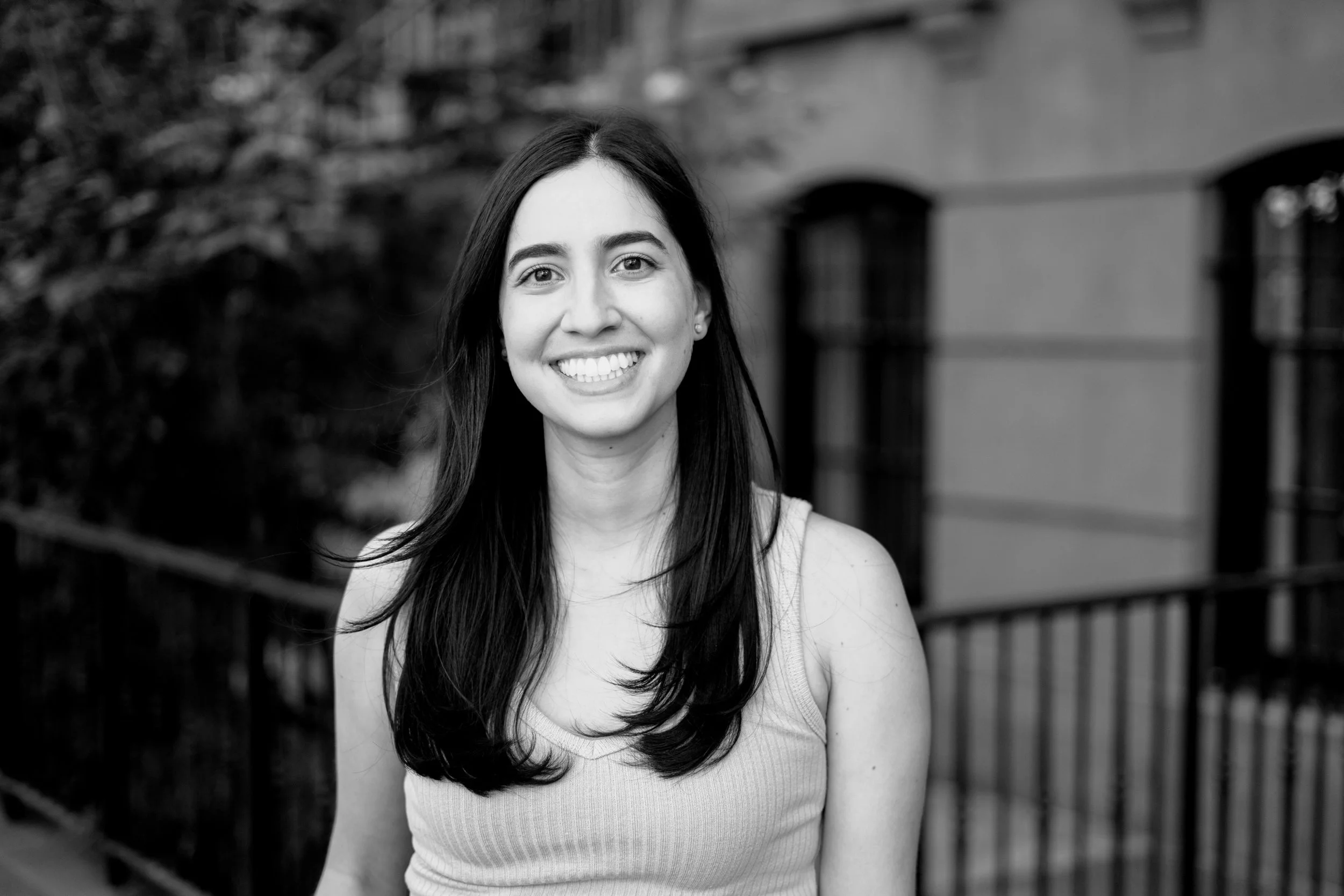 A black and white portrait of a New York City therapist with long dark hair, wearing a sleeveless top, standing outdoors in front of a building with arched windows.