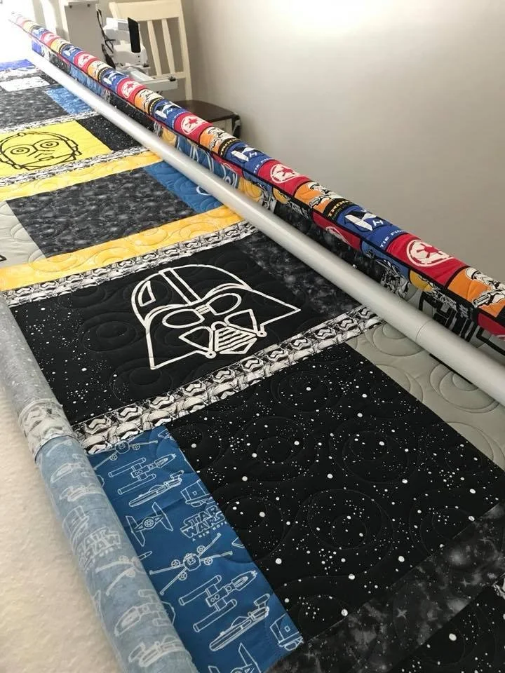 A quilt with Star Wars themed fabrics, featuring Darth Vader and space patterns, laid out on a bed with rolled-up Star Wars fabric borders.