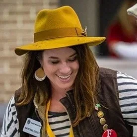 Woman smiling and wearing a yellow hat, striped shirt, and vest with buttons.