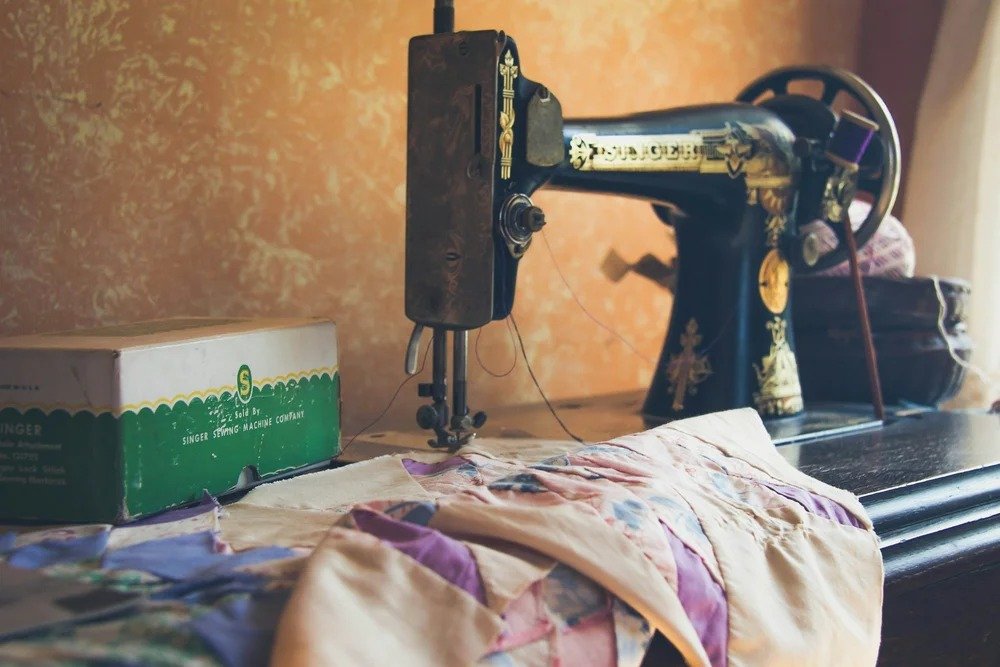 An antique black and gold sewing machine with ornate details on a wooden table, with a partially sewn colorful fabric in front and a green and white box labeled 'Singer' on the left, against a warm-toned wall.