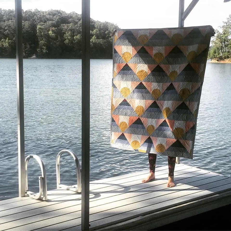 Person standing on a dock near water, holding a colorful quilt in front of their face, with only their legs visible.