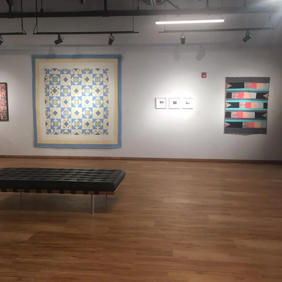 Art gallery with various quilts on display, wood flooring, gray walls, and track lighting.