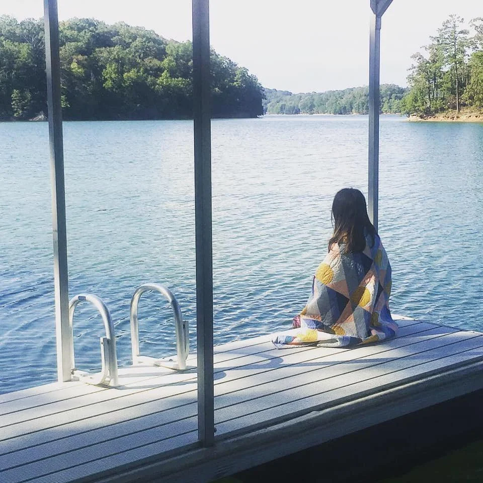 A woman wrapped in a colorful quilt sitting on a dock by a lake, looking out at the water and distant trees.