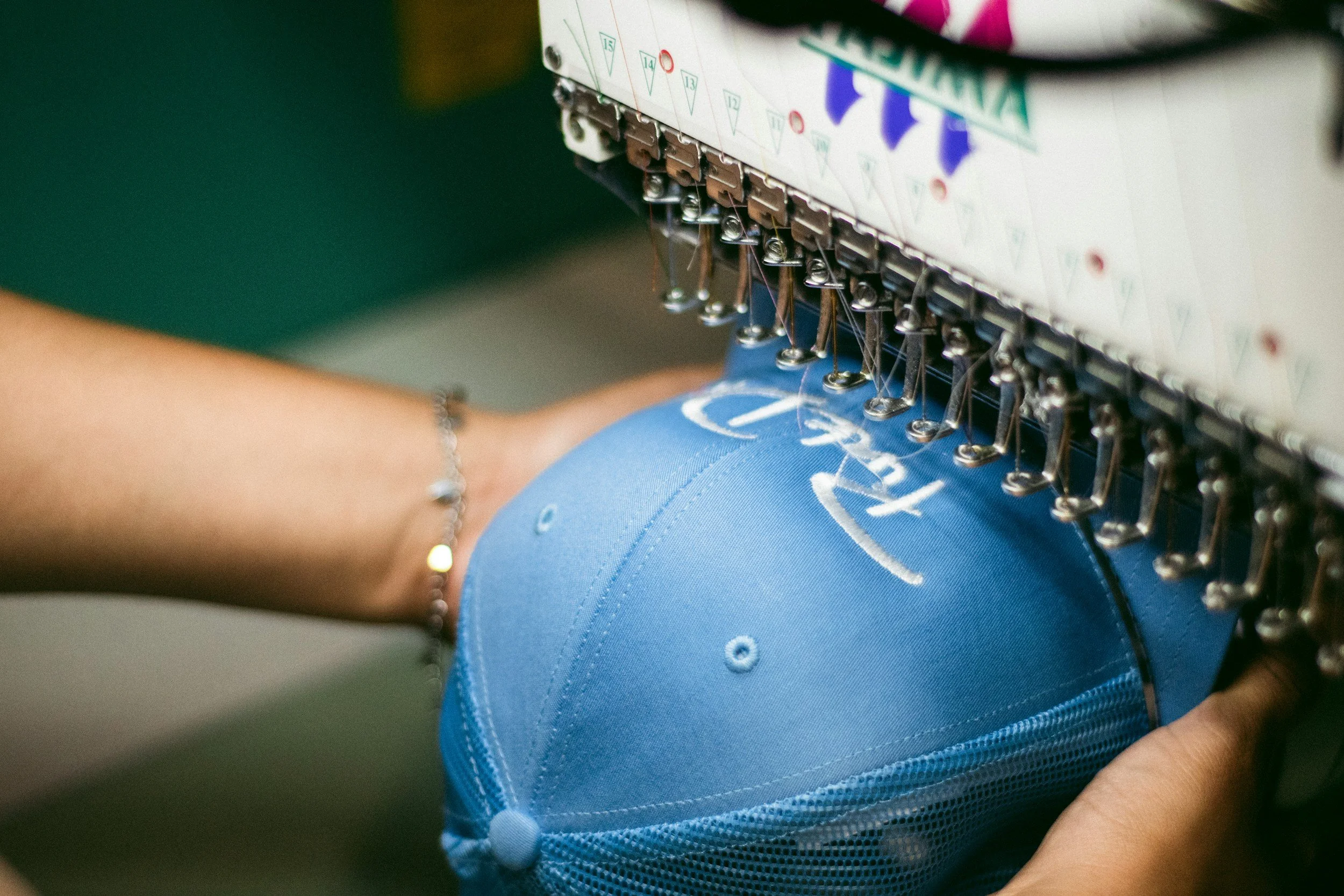 A person using an embroidery machine to sew white cursive text onto a blue baseball cap.