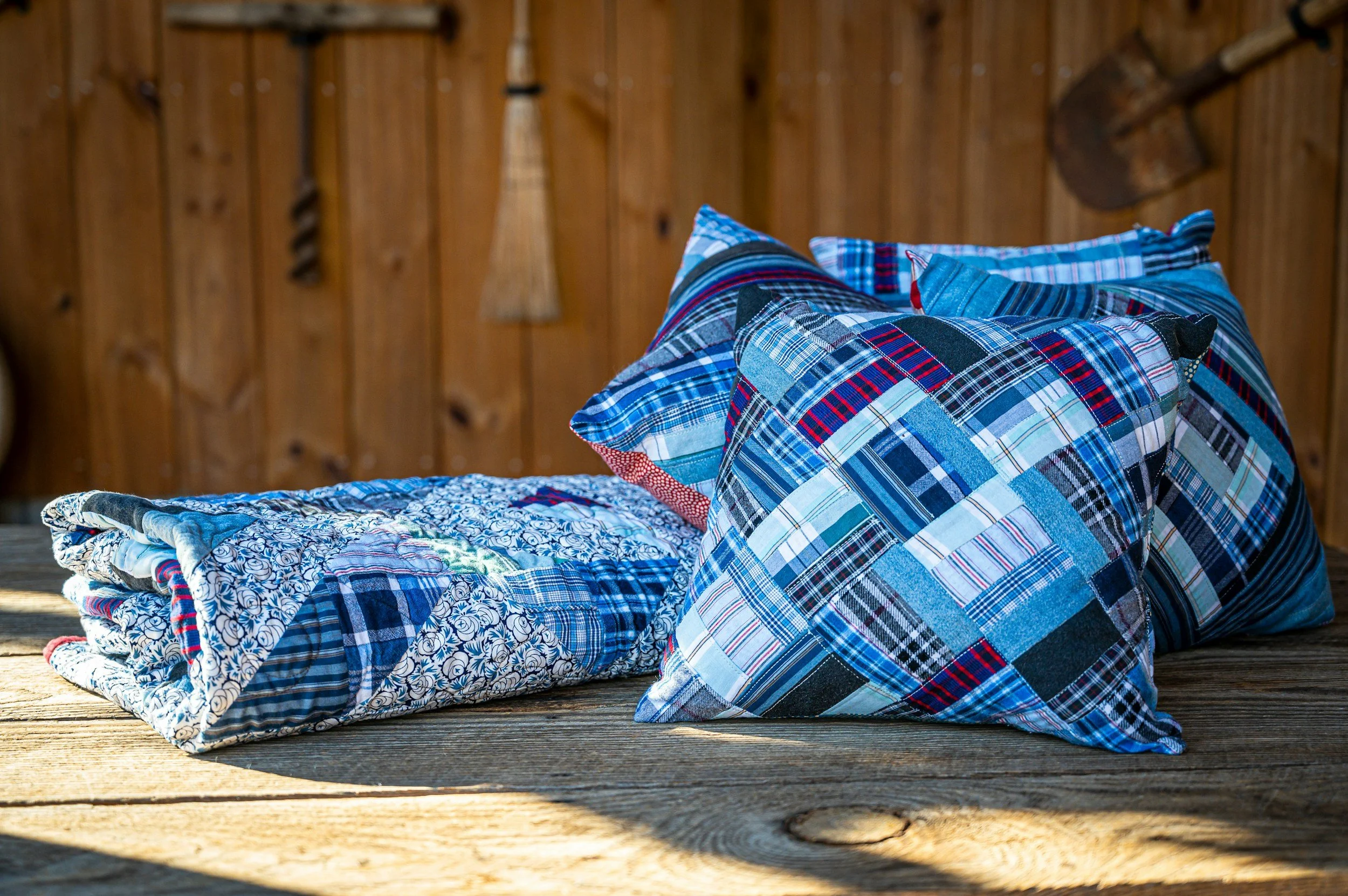 A stack of folded quilts and pillows made from blue and red plaid and floral fabrics, resting on a wooden surface in front of a wooden wall with hanging tools.