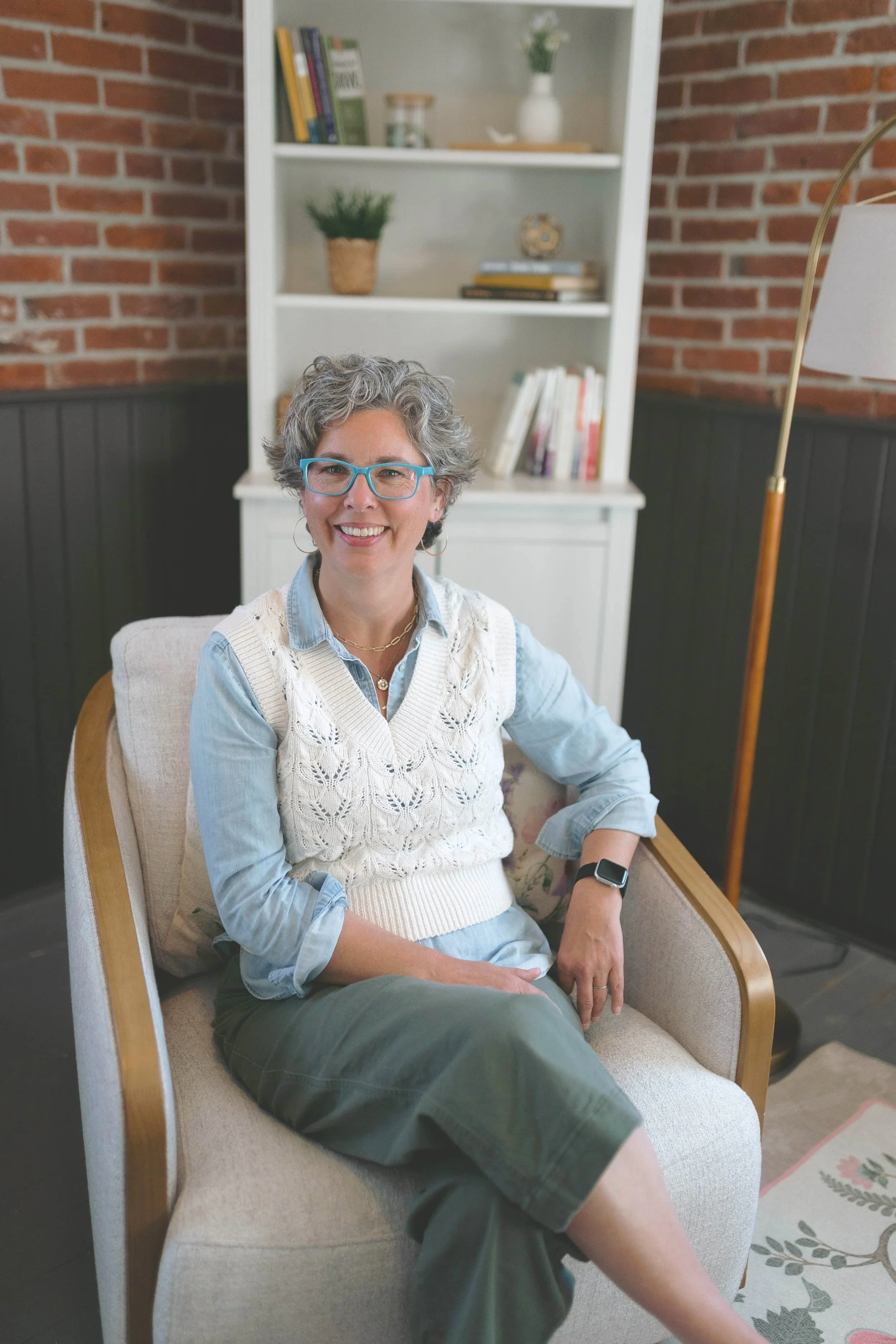 Kathleen Peters, grief therapist, smiling and seated in a cozy office armchair with a bookshelf and exposed brick wall in the background.