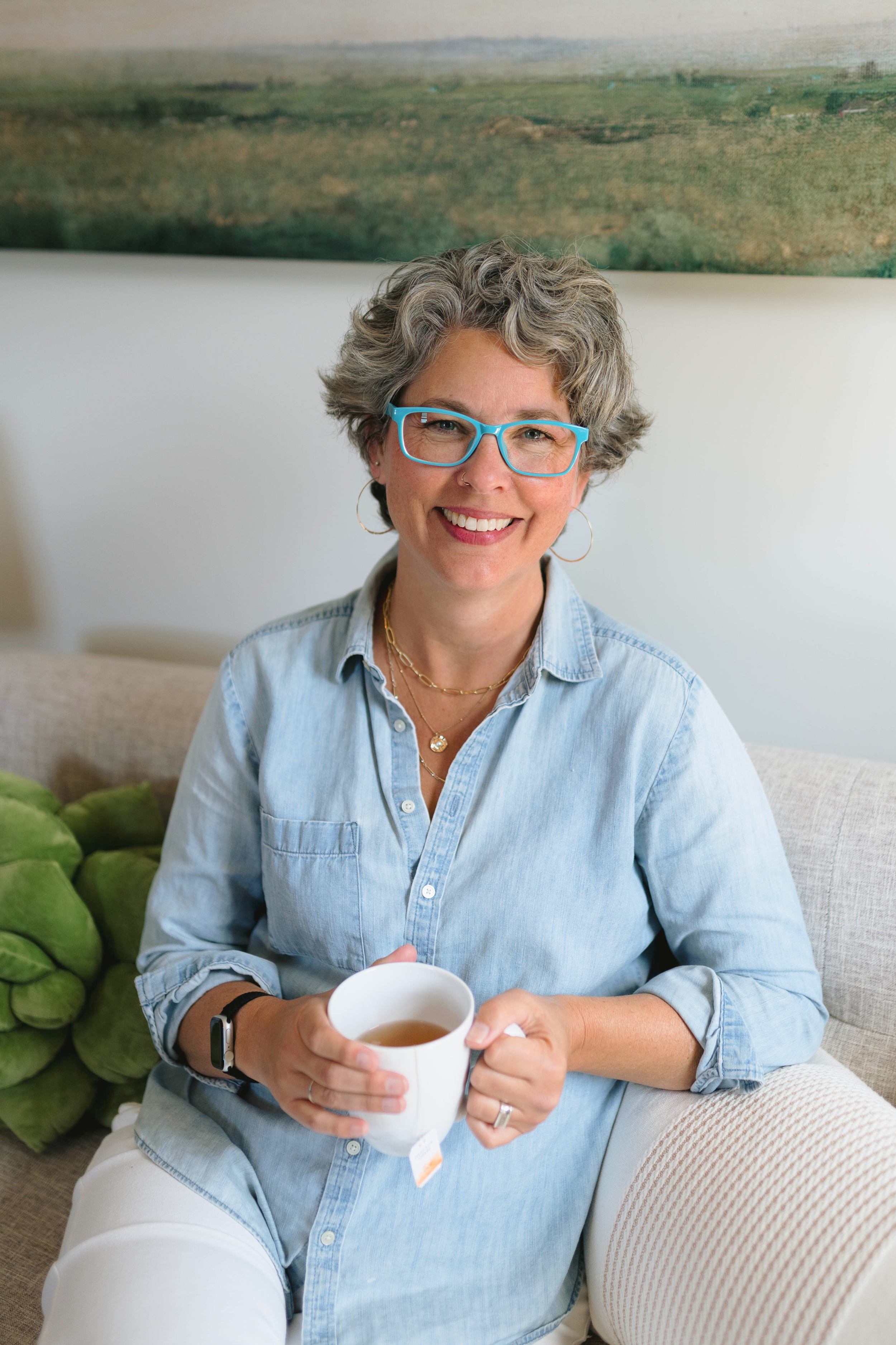 Kathleen Peters, grief therapist, sitting on a couch and smiling while holding a mug of tea.