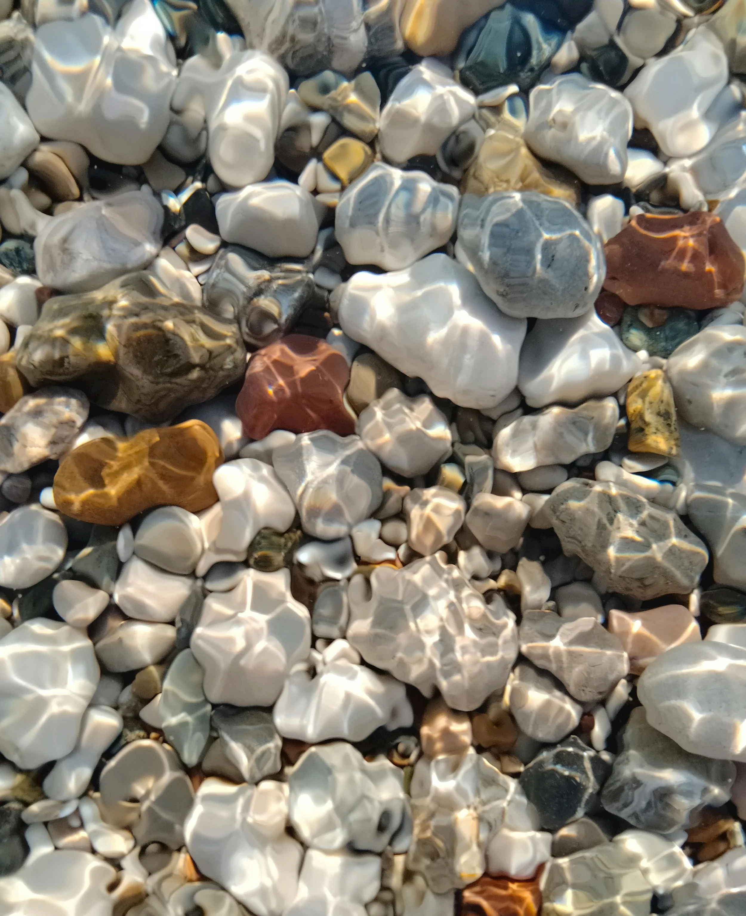 Close-up of small, smooth, multicolored pebbles on the beach, reflecting sunlight.