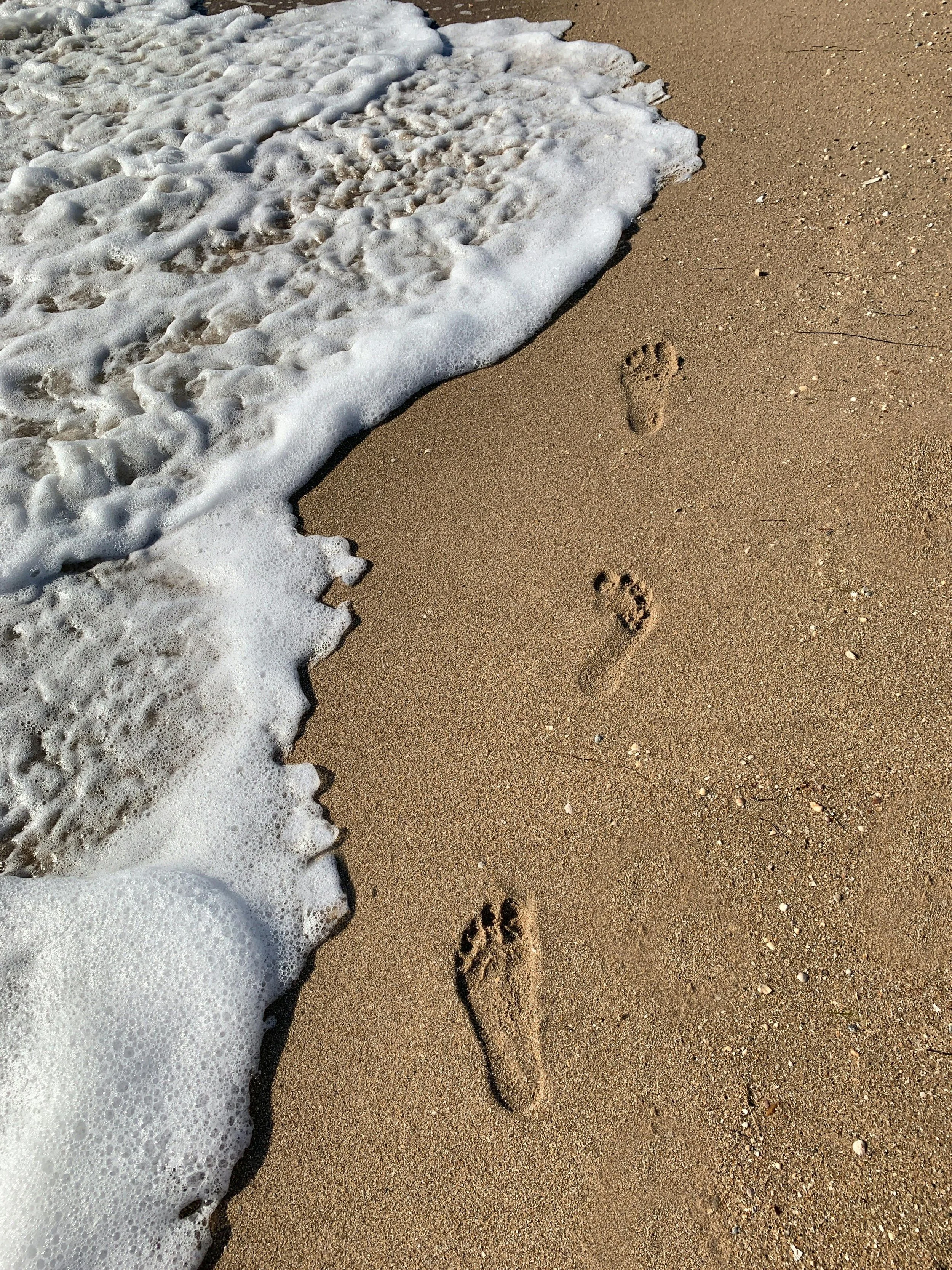 Footprints in the sand on a beach with foamy ocean waves approaching.
