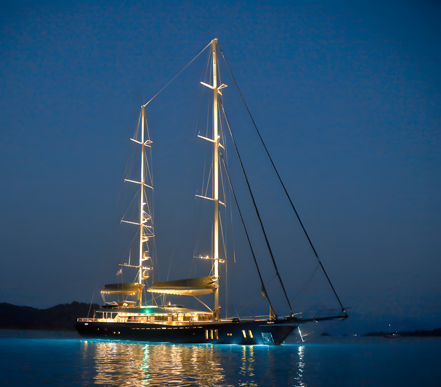 A luxury sailing yacht illuminated at night, floating on calm water with a dark sky and land in the background.