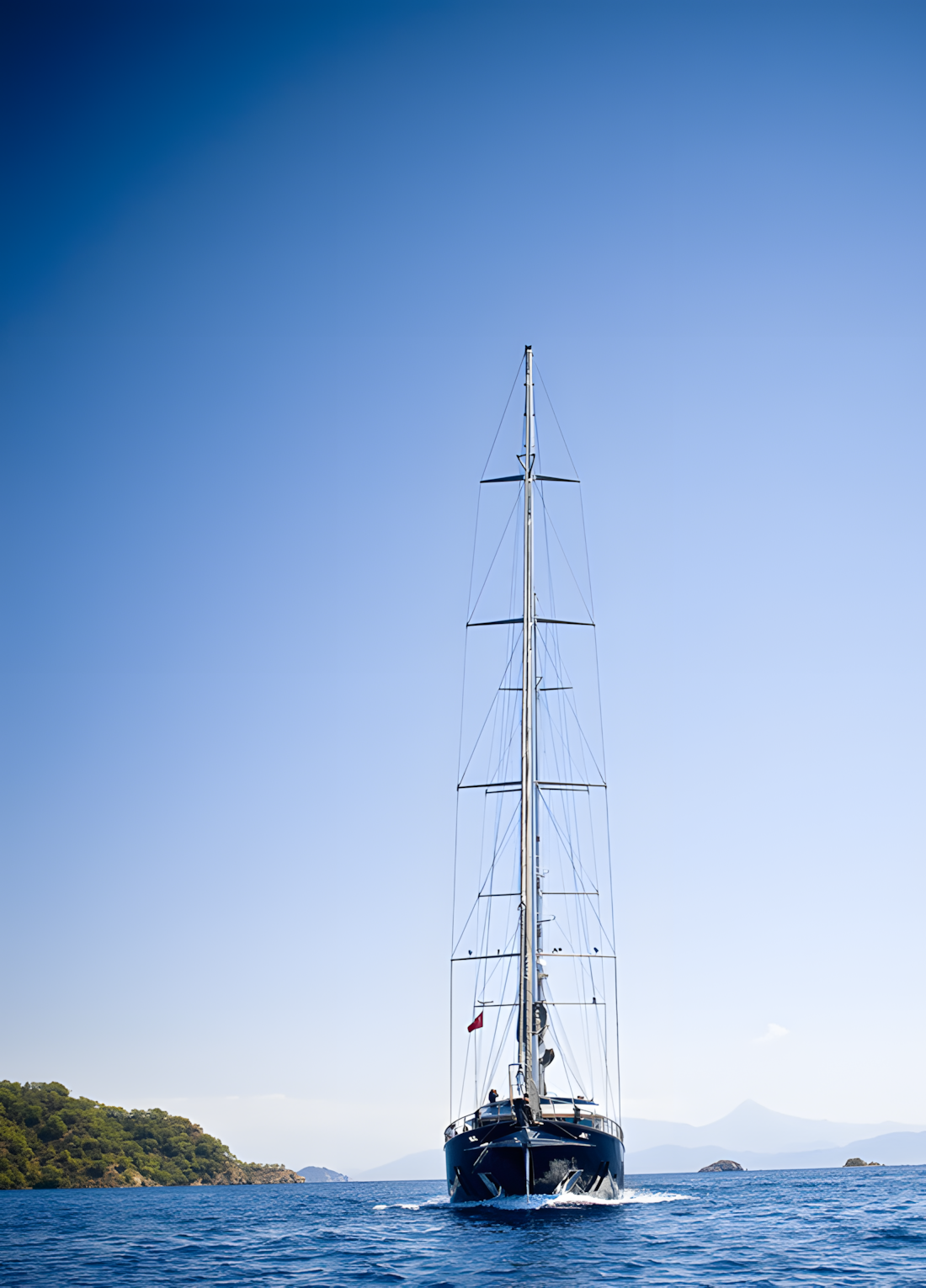 A large black sailboat with its sail down, sailing on calm blue waters with small islands and mountains in the background under a clear blue sky.