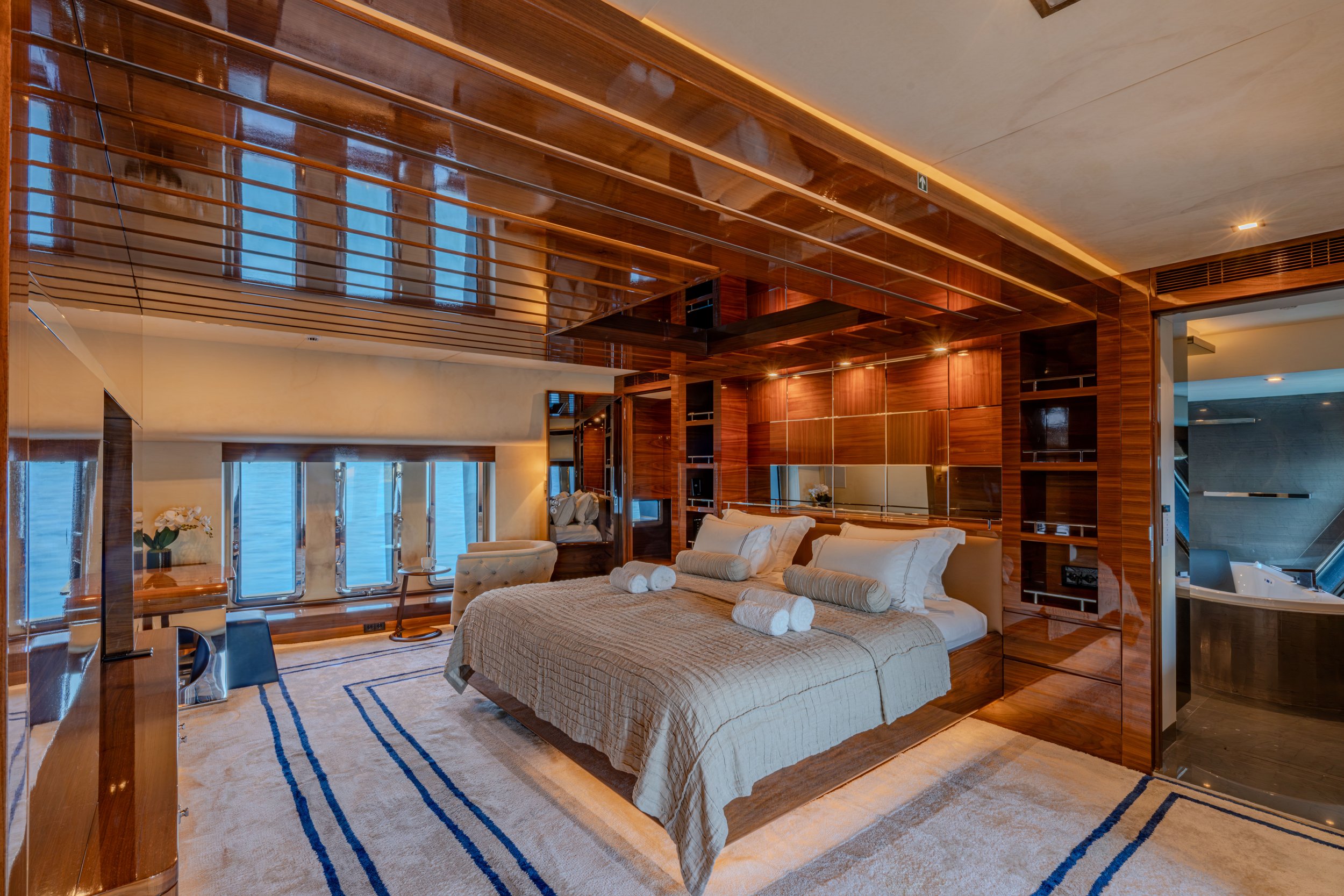 Luxury bedroom with large bed, wooden paneled walls, windows showing water view, and an en-suite bathroom with jacuzzi tub.