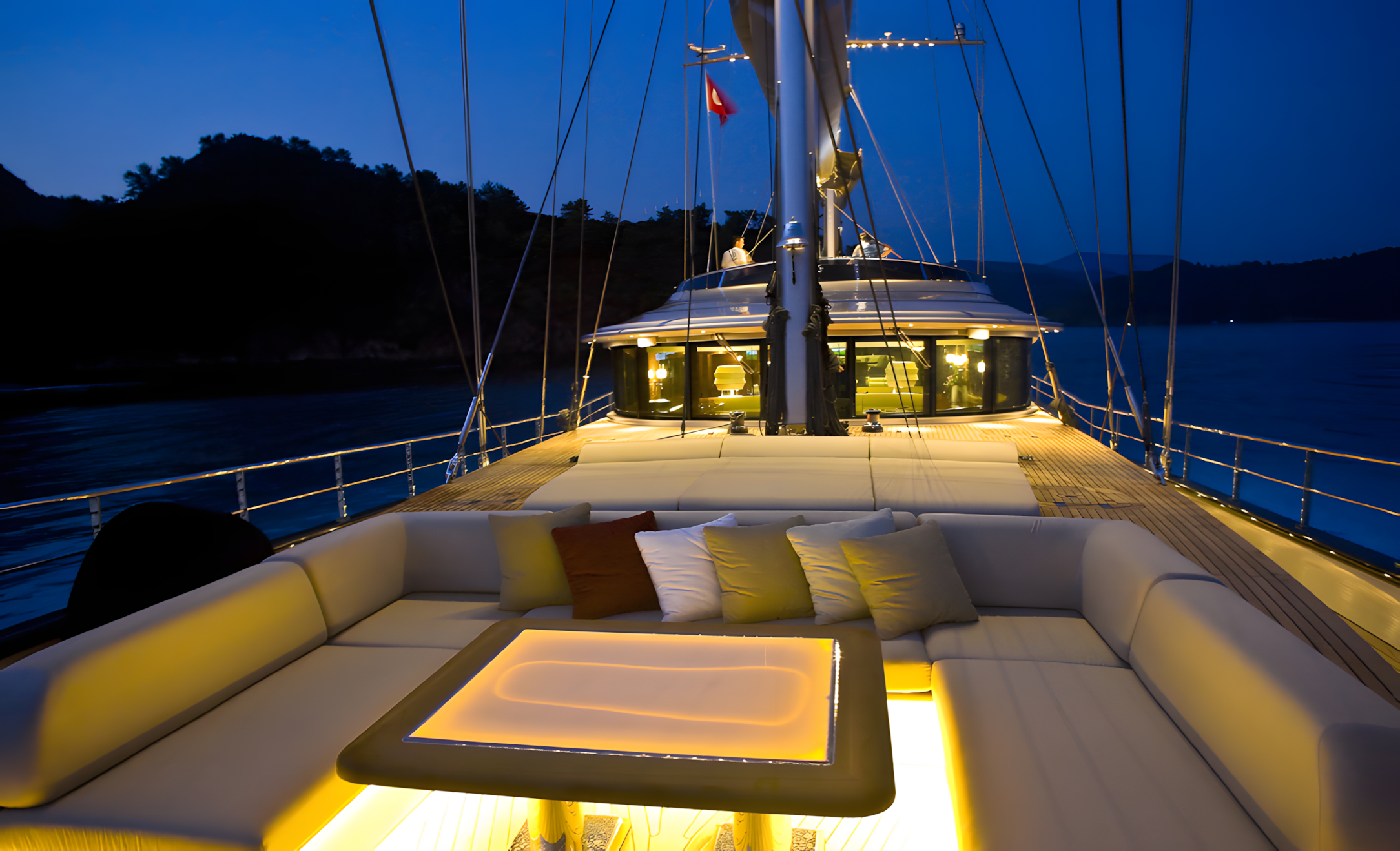  View of the deck on a yacht at night, illuminated with warm lighting. There are multiple cushions on the seating area, and a rectangular illuminated table in the center. In the background, the yacht's bridge and mast are visible, with some people on