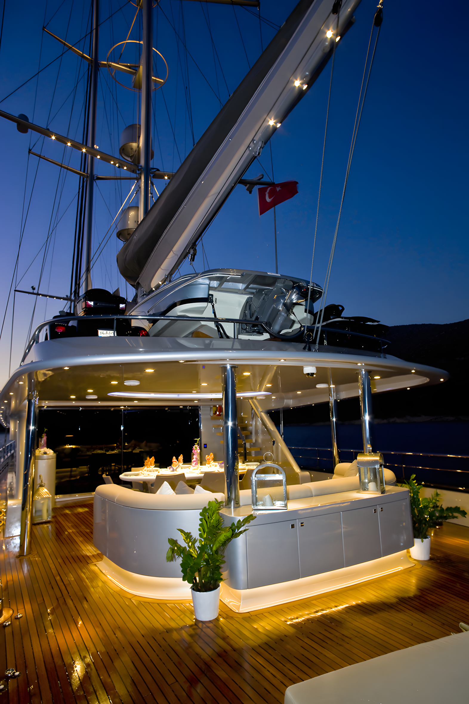 A luxury yacht at night, illuminated with interior and exterior lighting, featuring a dining area with a table set for a meal, potted plants, and a Turkish flag in the background.