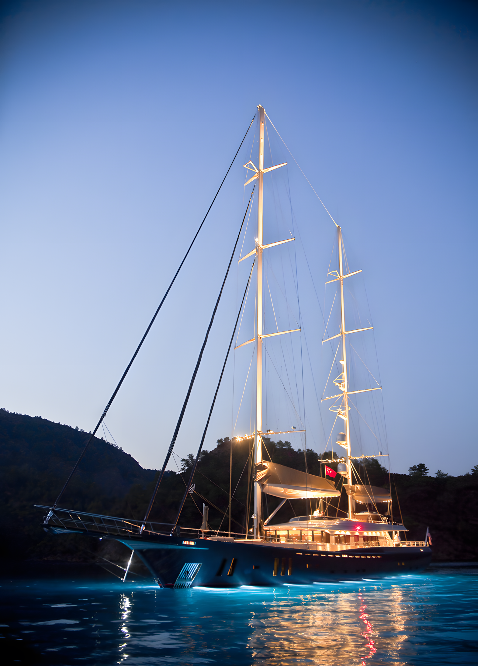 A sleek black sailing yacht illuminated with lights, floating on calm water during dusk with a dark hillside in the background.