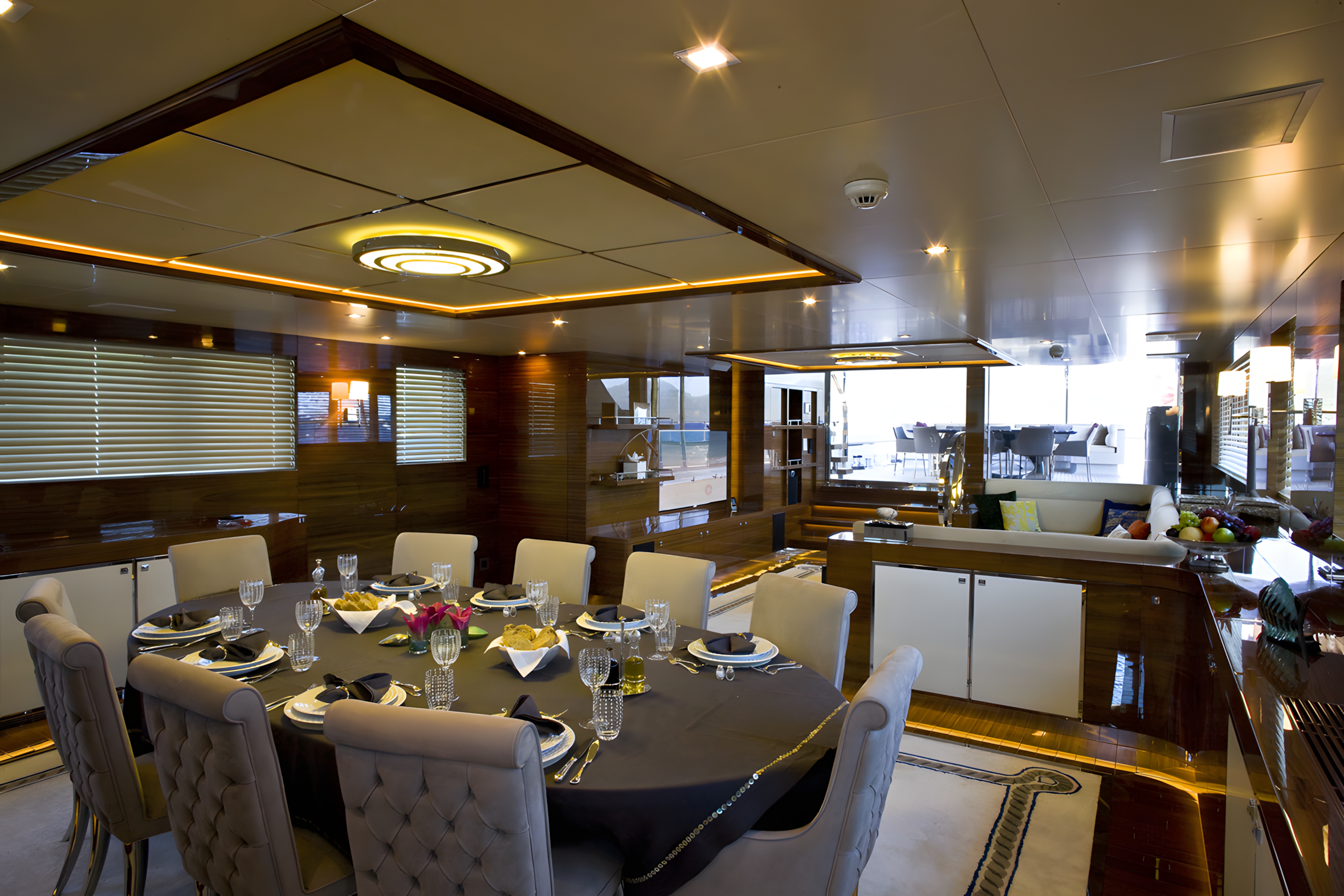 Elegant dining area with a round table set with black napkins, glassware, and a centerpiece of pink flowers, in a luxury yacht with wooden walls and large windows.