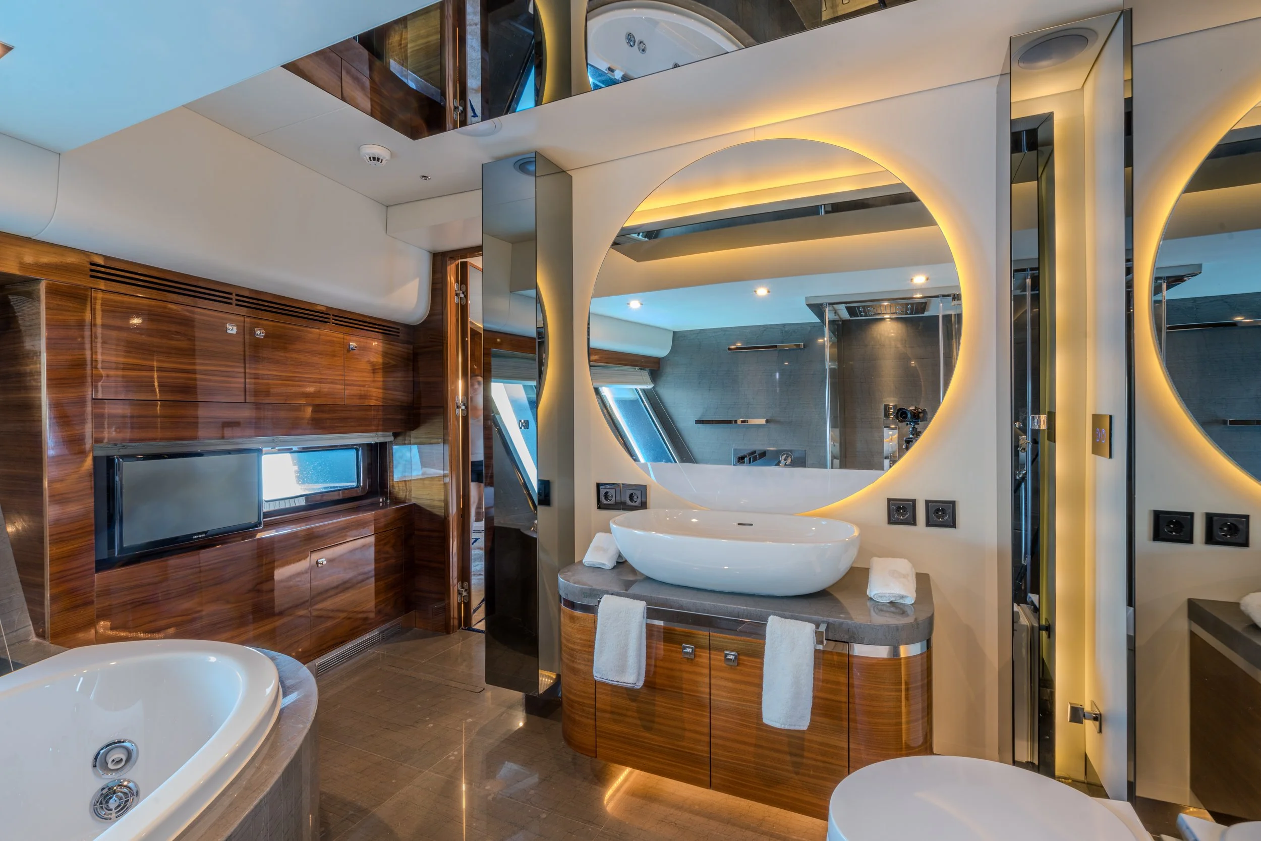 Modern bathroom with wooden cabinetry, large mirror with backlighting, white vessel sink, and a whirlpool bathtub visible in the foreground.