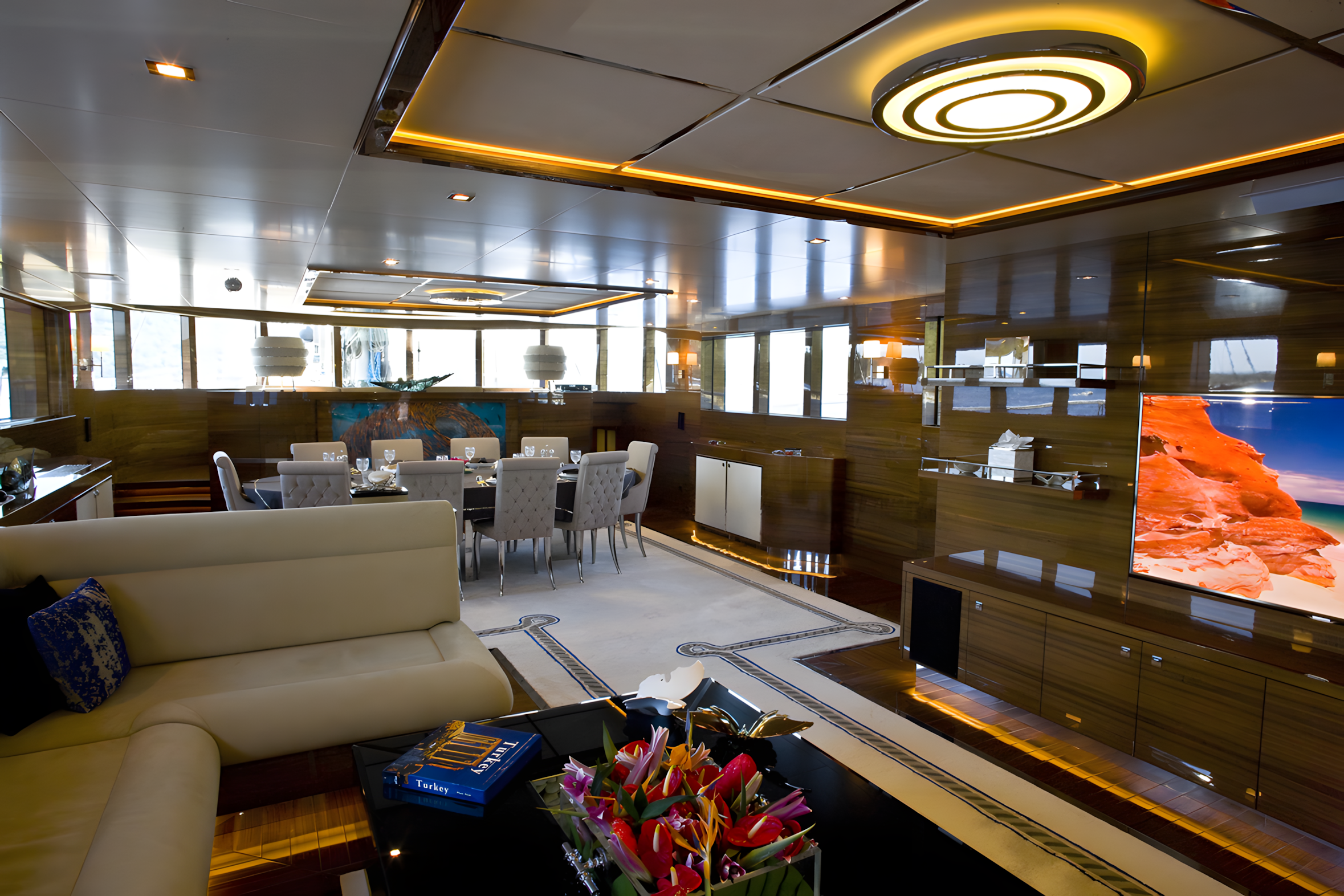 Luxury yacht lounge with wooden paneling, white leather sofa, dining table with chairs set for a meal, modern lighting, large flat-screen TV, floral centerpiece, and large windows with a view of the sea.