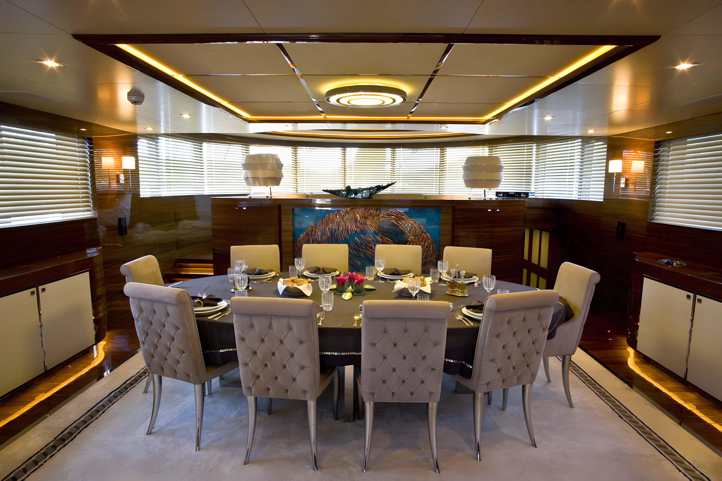 Elegant dining room with a round black table set for eight, surrounded by tan tufted chairs. The table is decorated with a pink floral centerpiece and drink glasses, with napkins and plates set for a formal meal. The room features wood-paneled walls,