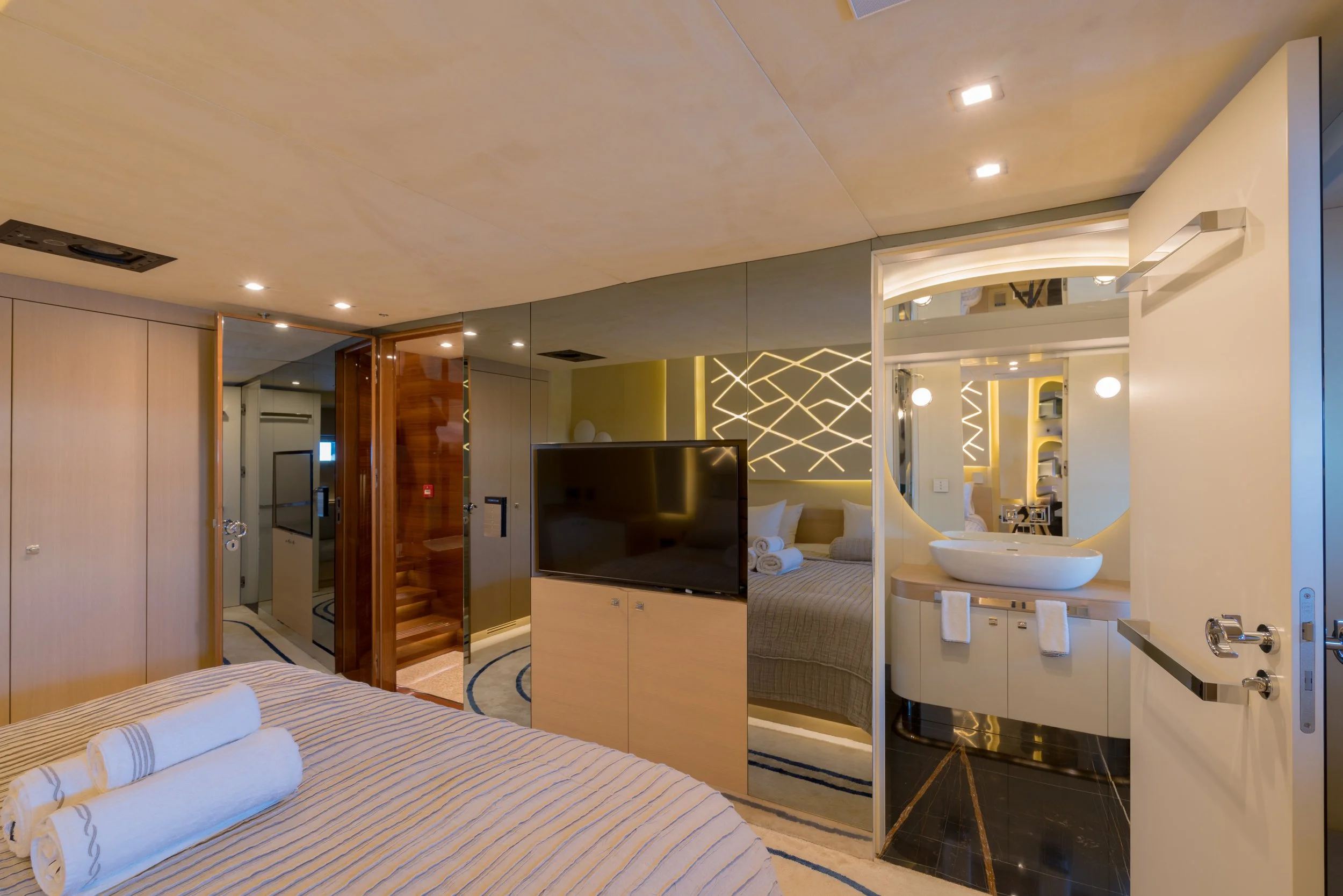 Luxury hotel room with bed, towels, flat-screen TV, mirrored wall, and an open bathroom with sink and decorative lighting.