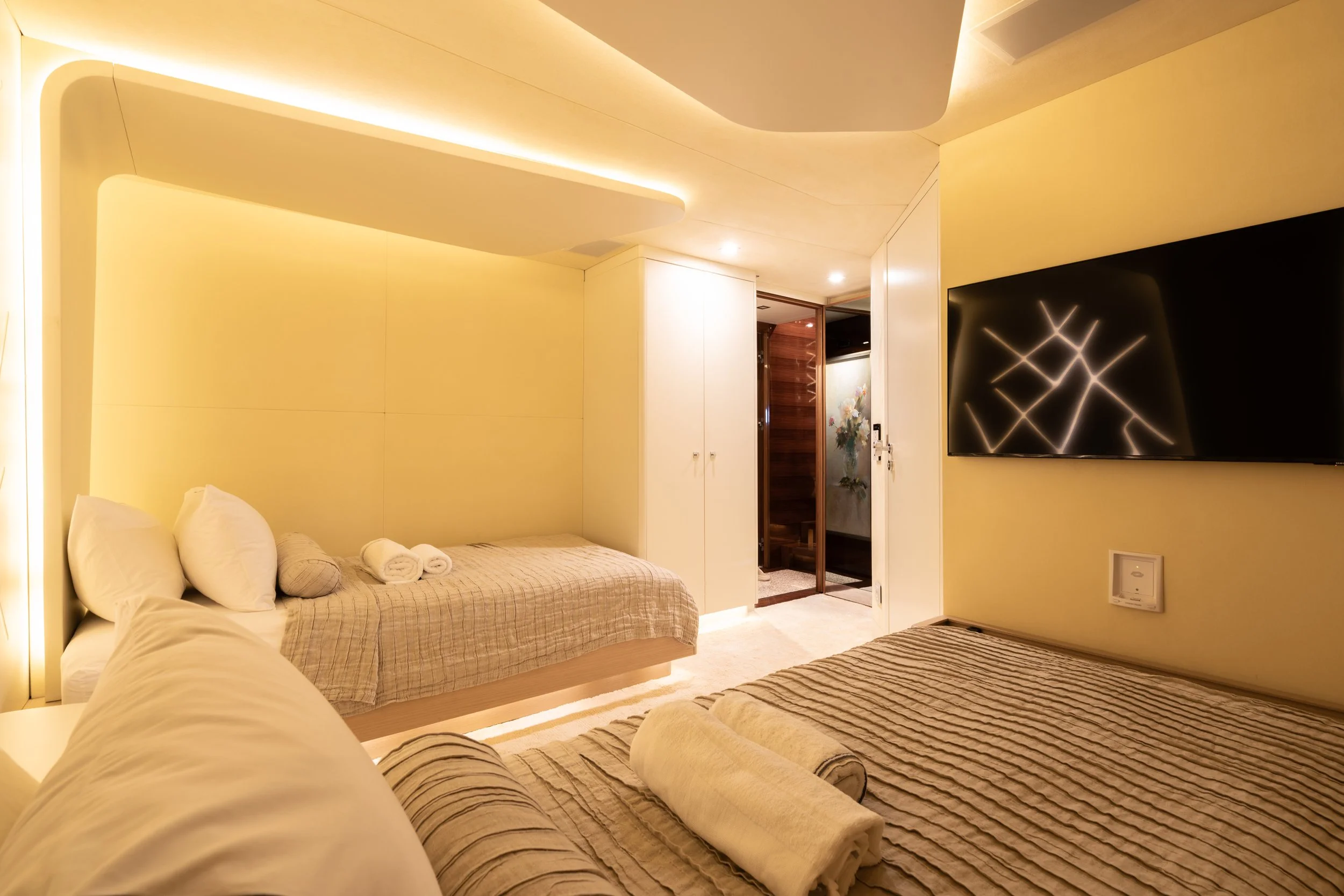 A hotel room with two beds, one in the foreground and one in the background, both with beige and striped linens. There are rolled towels on each bed. A large flat-screen TV is mounted on the wall opposite the beds. The room has warm lighting and a do